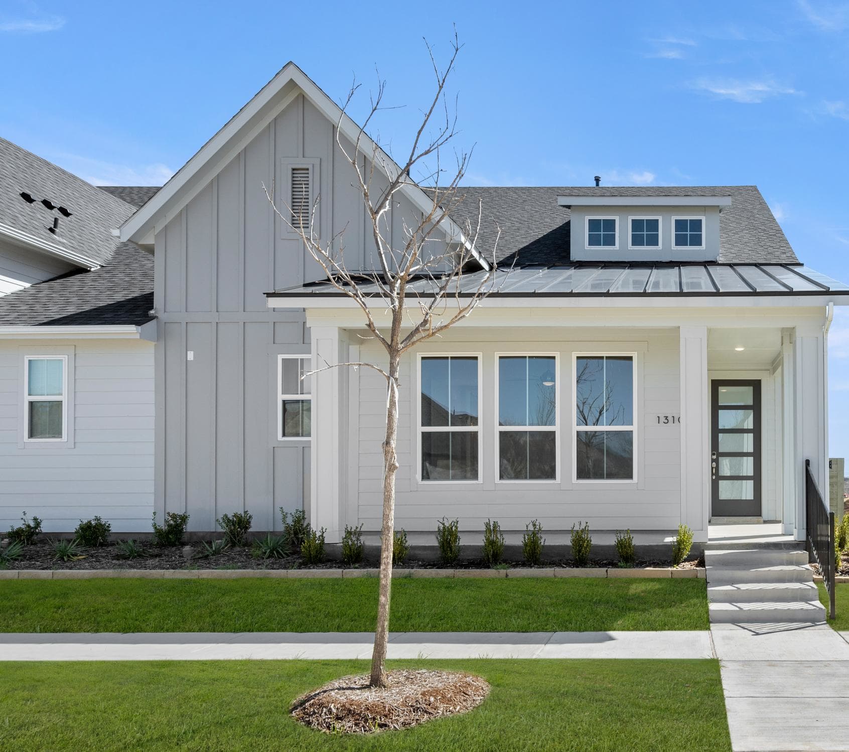 A modern, two-story house with a gray exterior, a metal roof, and a bare tree in the front yard, set against a clear blue sky.