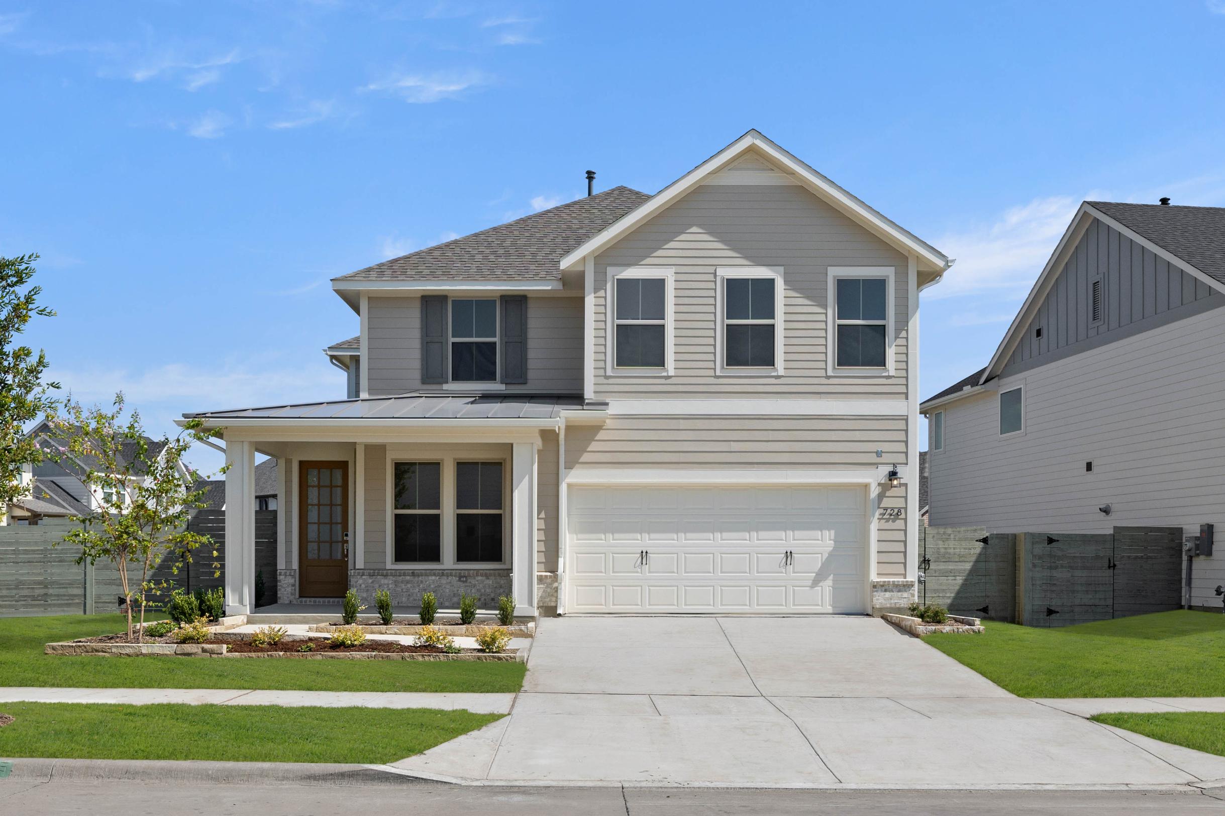 A two-story beige house with a gabled roof, surrounded by a well-manicured lawn and landscaping, stands against a clear blue sky.