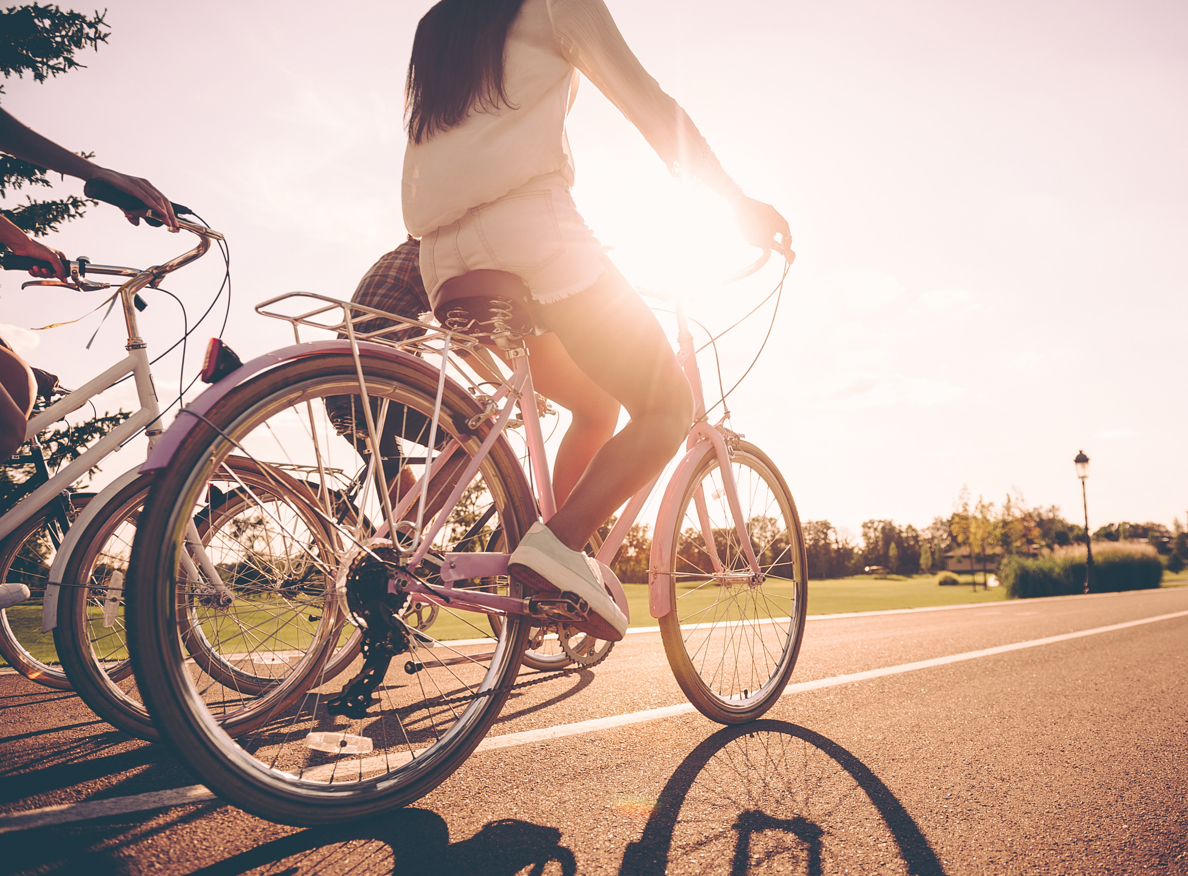A person riding a bicycle on a paved path surrounded by trees and a park-like setting, with the sun's rays creating a warm, backlit effect.