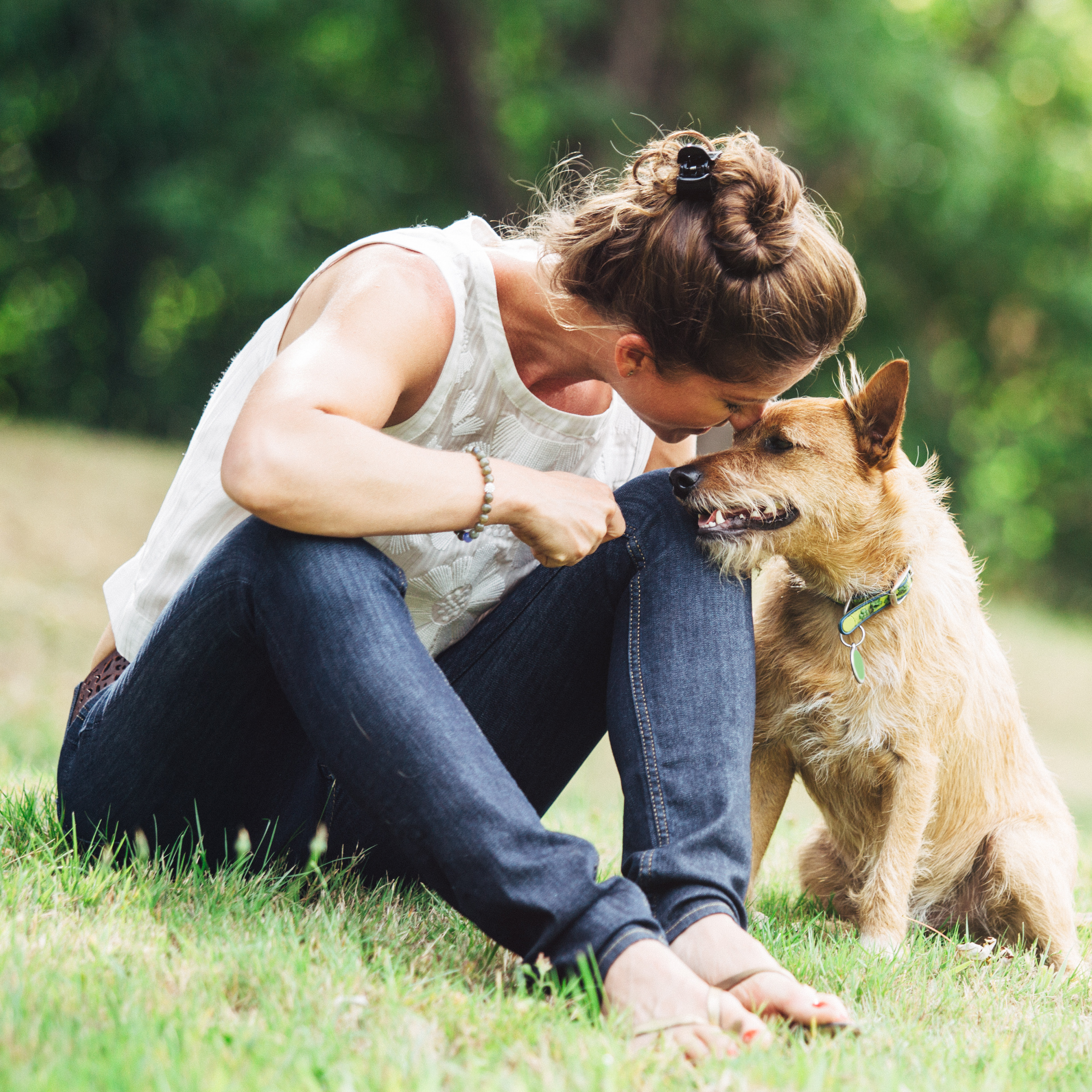 A woman in a white tank top is sitting on the grass, embracing a German shepherd dog in a lush, green outdoor setting.