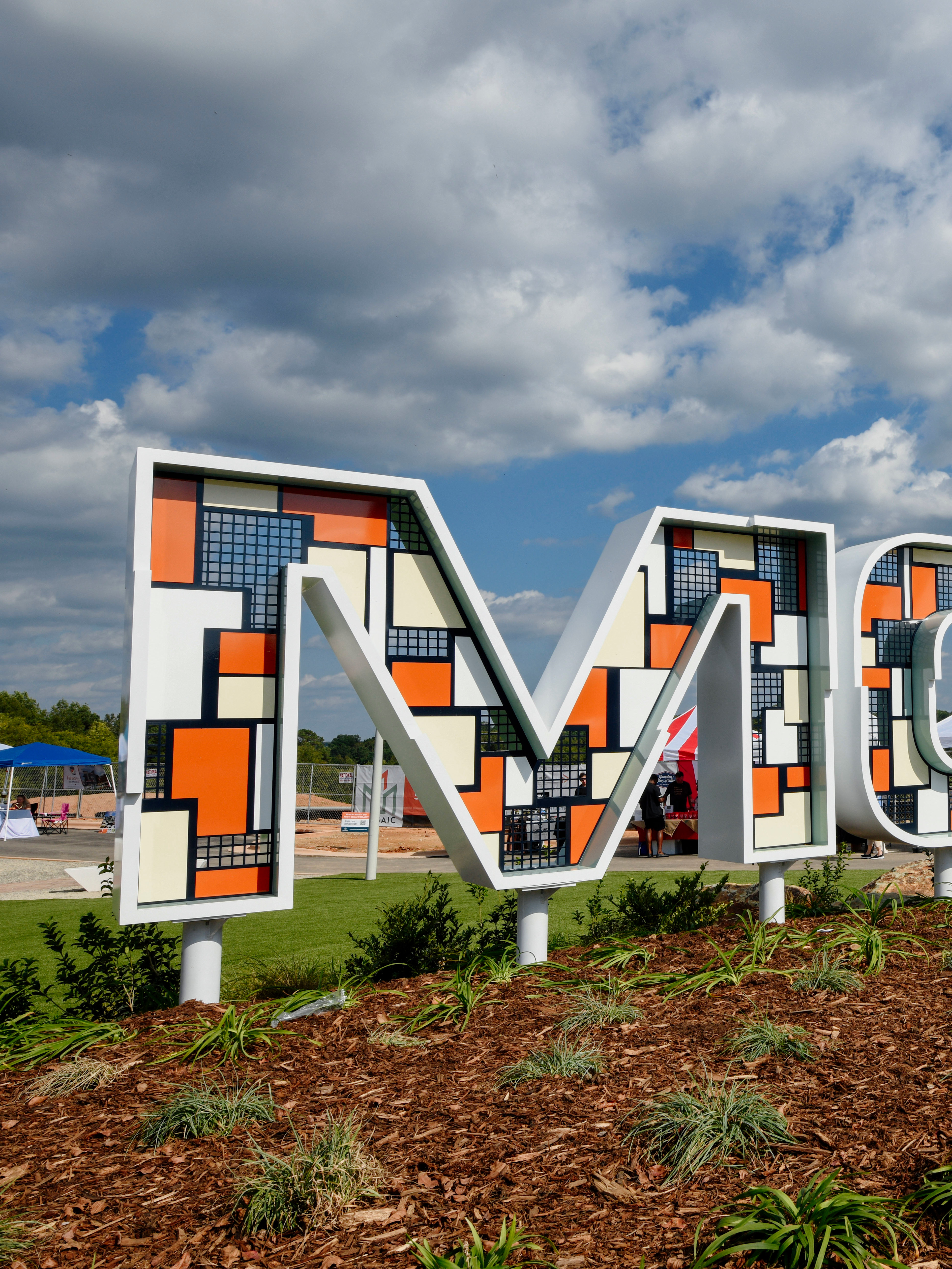 The image depicts a large, colorful sign spelling out "MOSAIC" against a backdrop of cloudy blue skies, with lush greenery in the foreground.