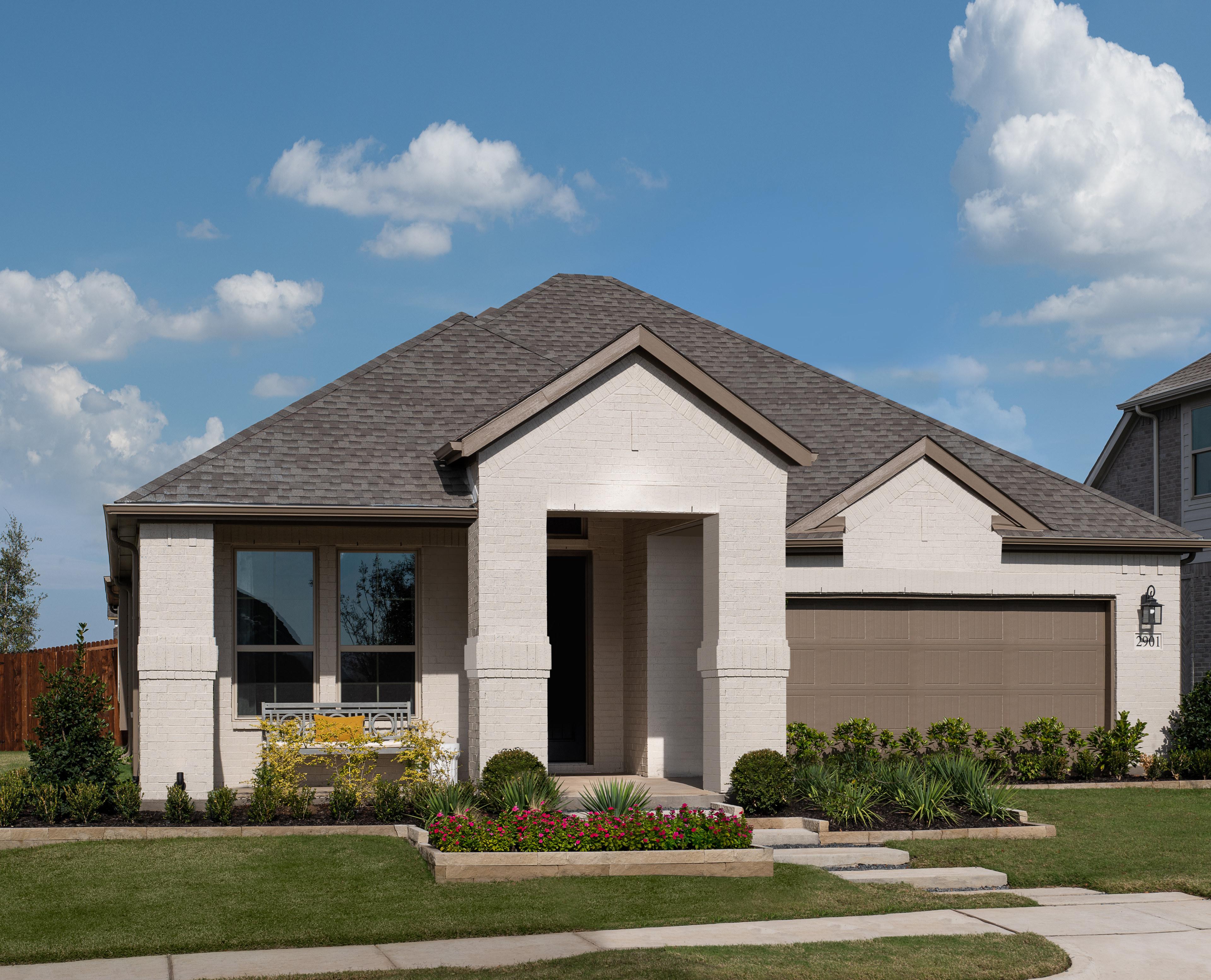 A well-manicured lawn surrounds a two-story house with a gabled roof, stone and stucco exterior, and a covered porch leading to the entrance. The sky is clear with fluffy white clouds, creating a picturesque suburban scene.