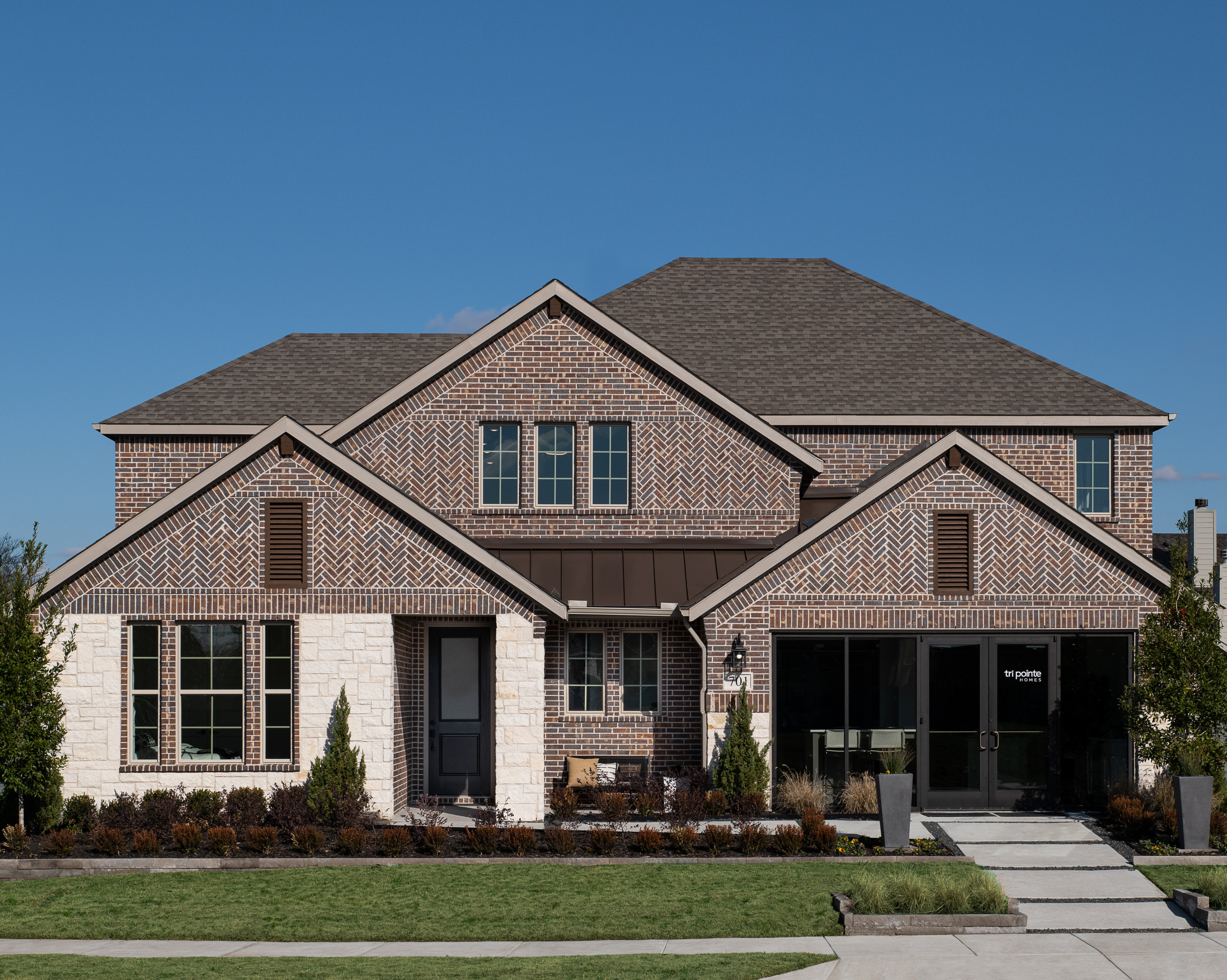 A two-story brick house with a gabled roof, surrounded by landscaped gardens and a paved driveway, set against a clear blue sky.