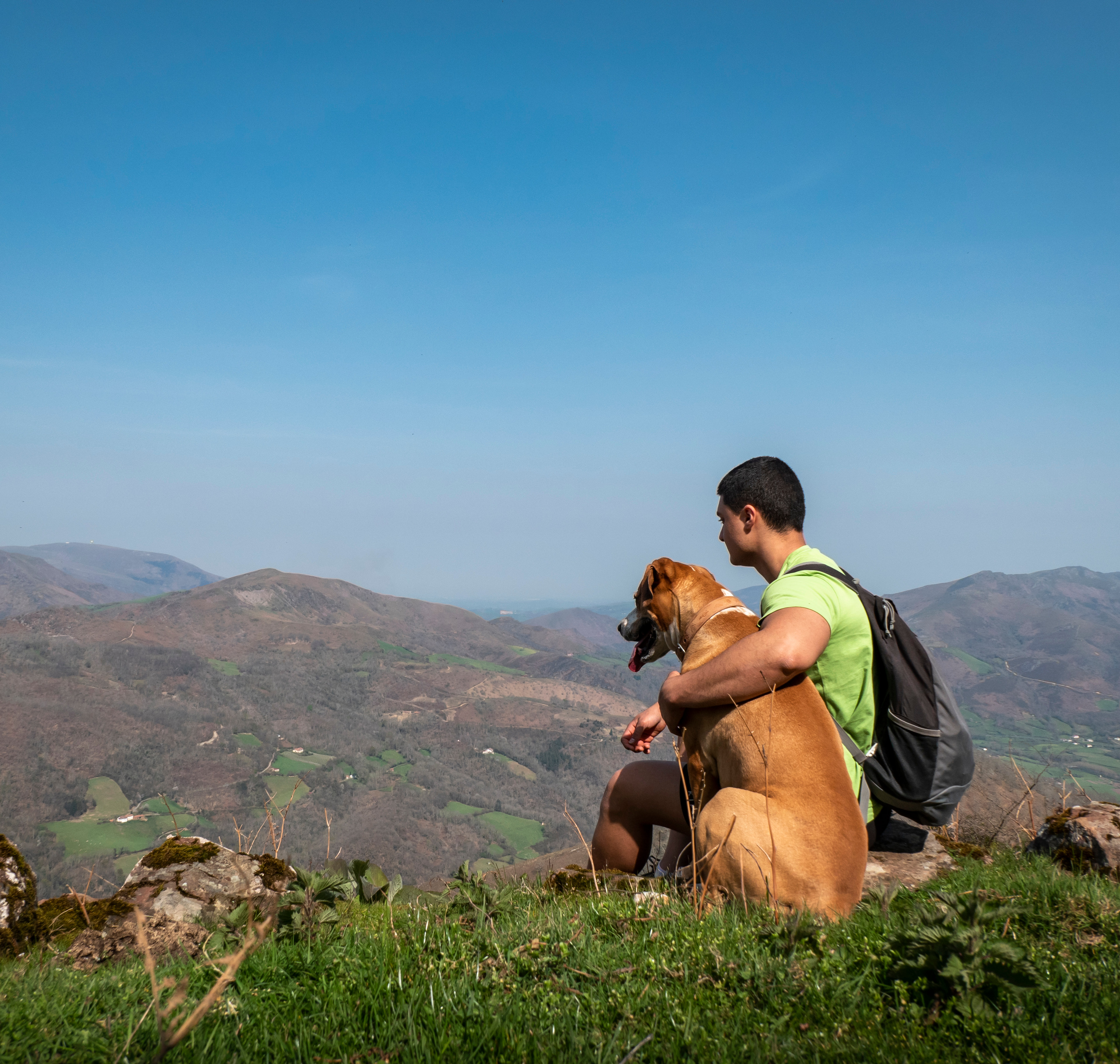 A person wearing a backpack stands on a grassy hill, overlooking a mountainous landscape in the background.