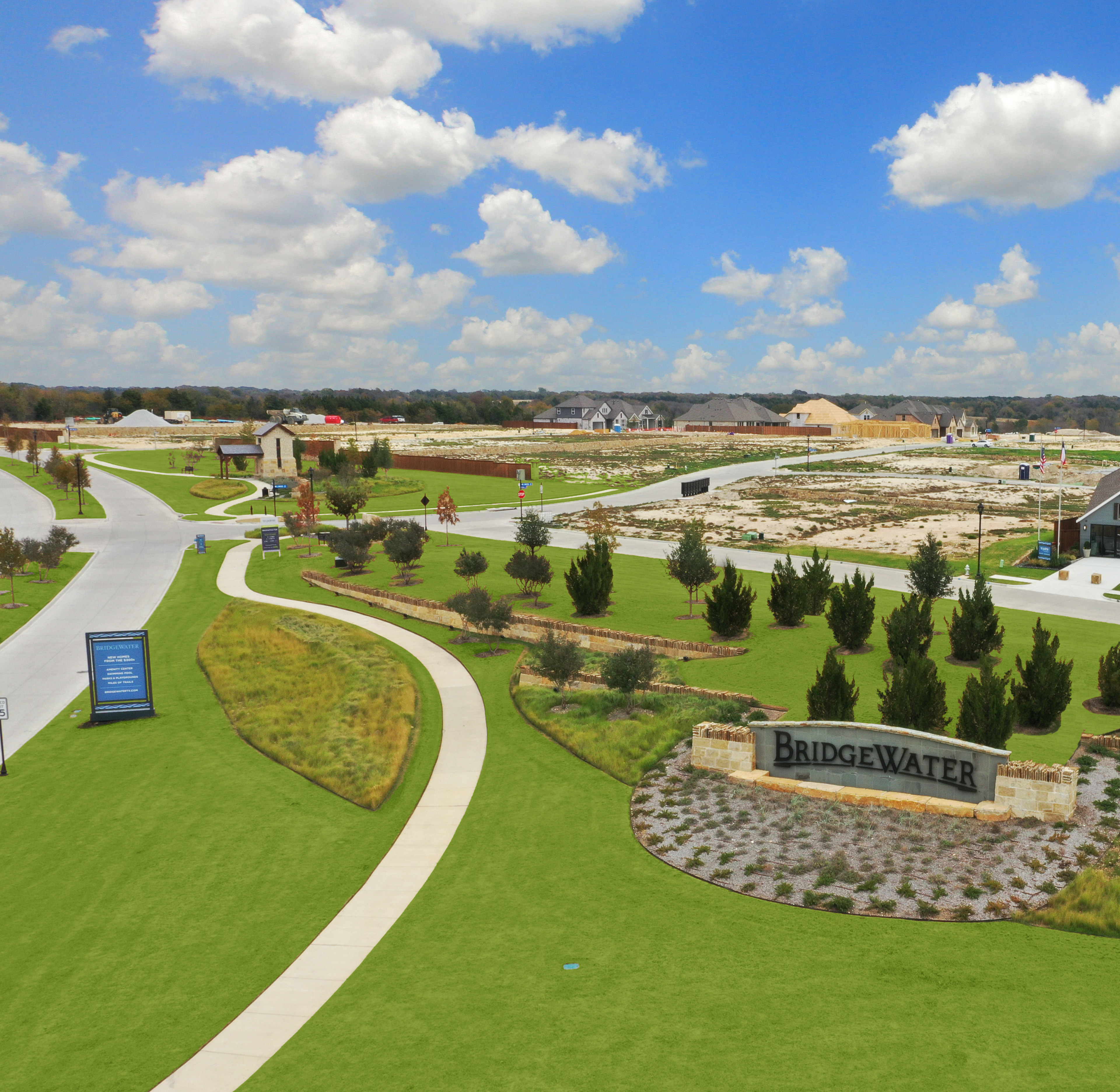 A well-manicured residential community with lush green lawns, neatly landscaped gardens, and a winding path leading through the development, set against a backdrop of a cloudy blue sky.