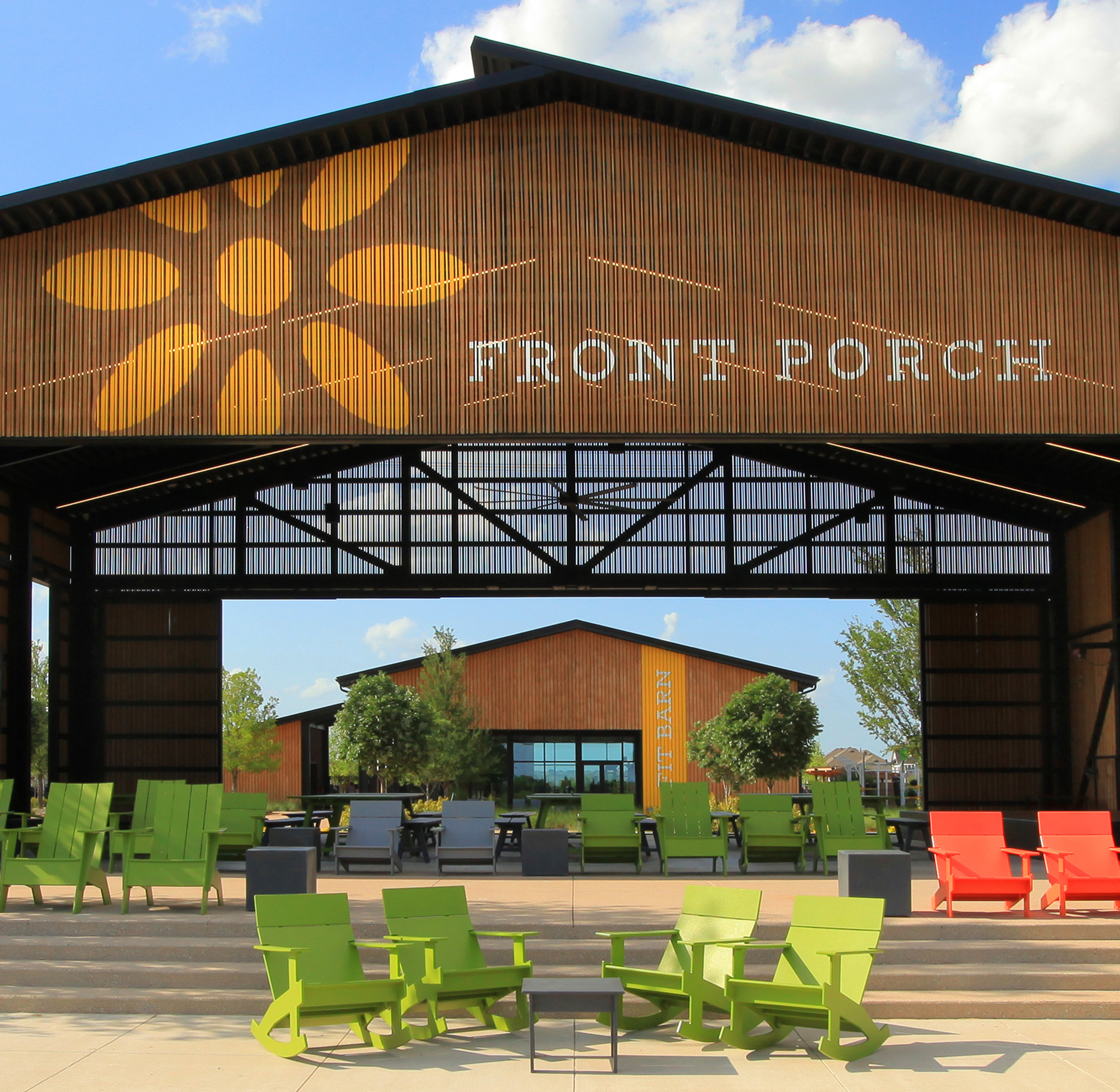 A wooden building with a large front porch, surrounded by colorful chairs and a grassy area under a blue sky with clouds.