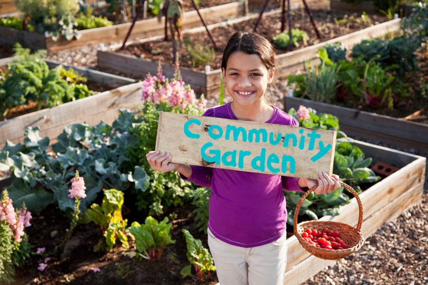 A smiling woman in a purple shirt holds a sign that says "Community Garden" while standing in a lush, vegetable-filled garden.