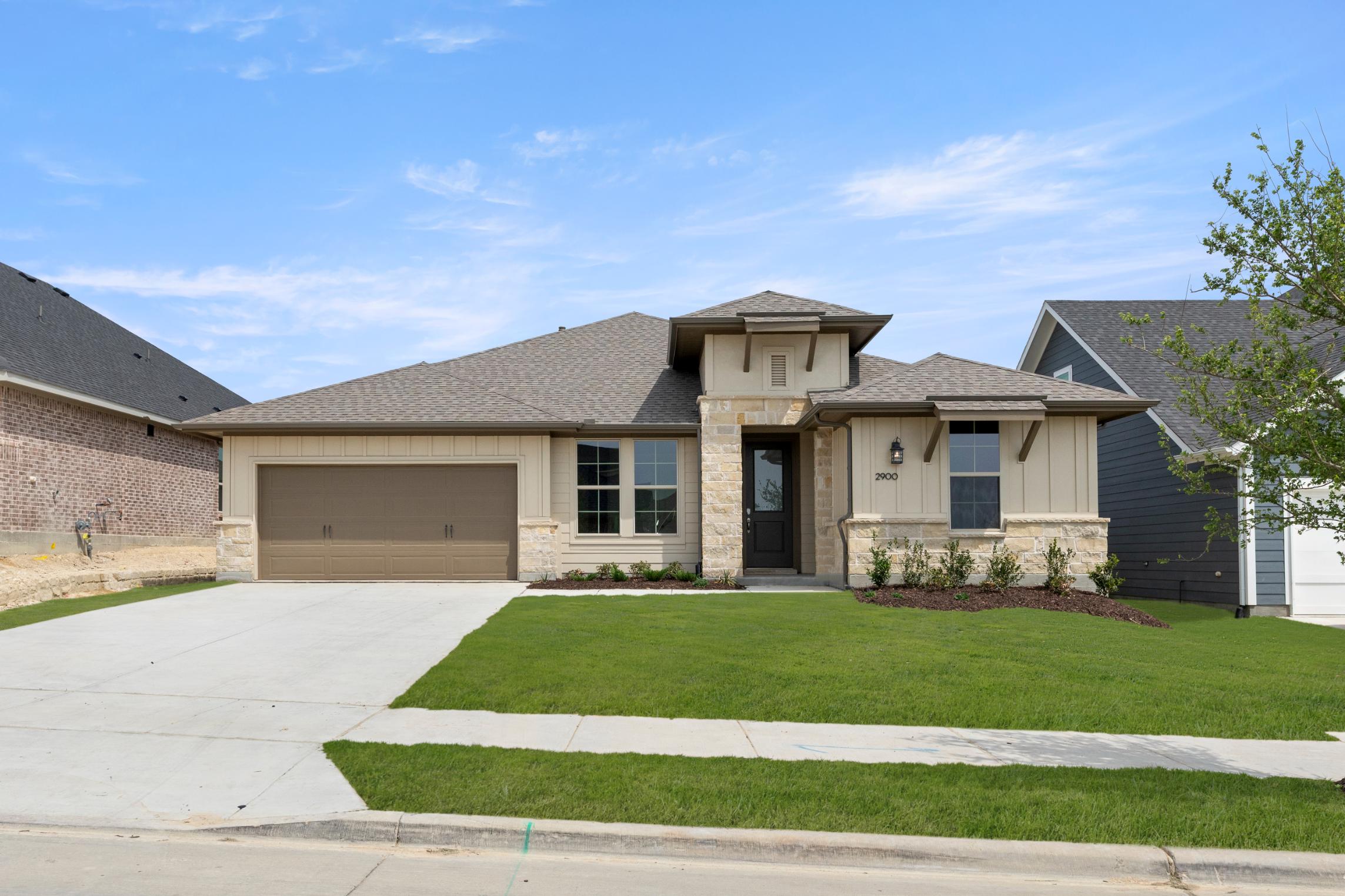 A well-manicured lawn surrounds a two-story house with a garage, featuring a mix of neutral and earthy tones in its exterior design, set against a clear blue sky with scattered clouds.