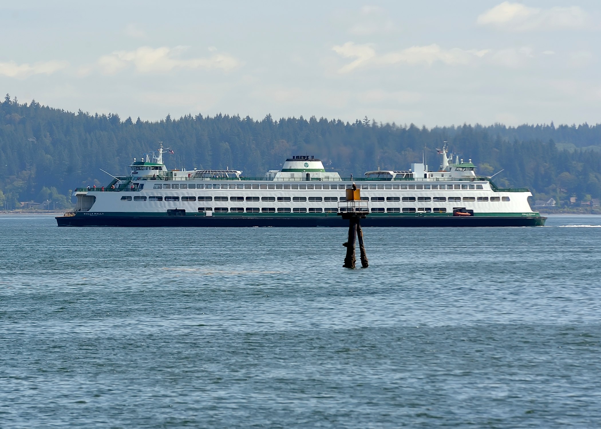 A large ferry boat is seen in the foreground, floating on the calm waters of a body of water surrounded by a forested landscape in the background.