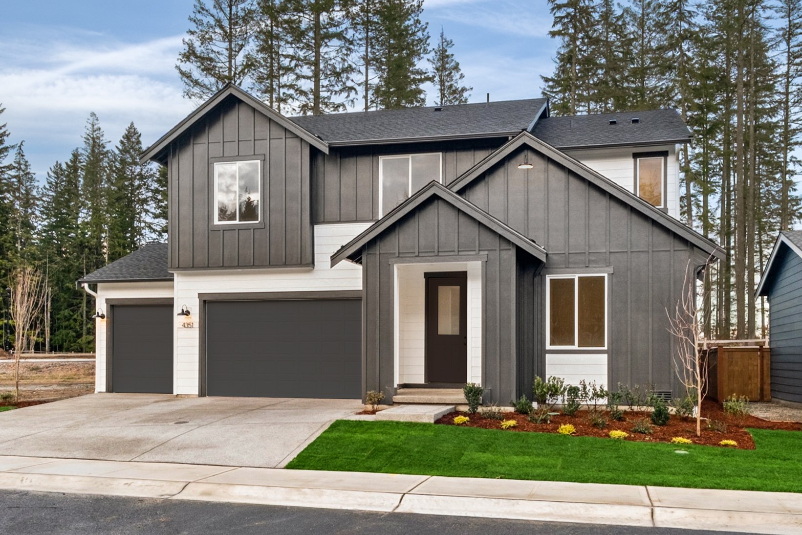 A modern two-story house with a gray exterior, a garage, and a well-manicured lawn with colorful flowers in the foreground, surrounded by a forested area in the background.