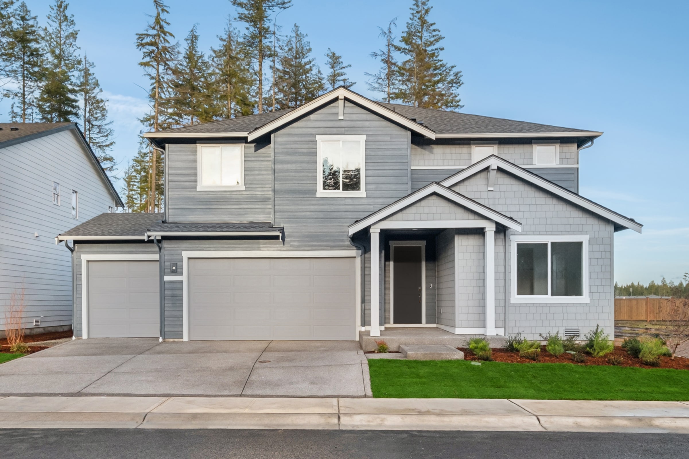 A two-story gray house with a garage stands in the foreground, surrounded by pine trees and a grassy lawn in the background.