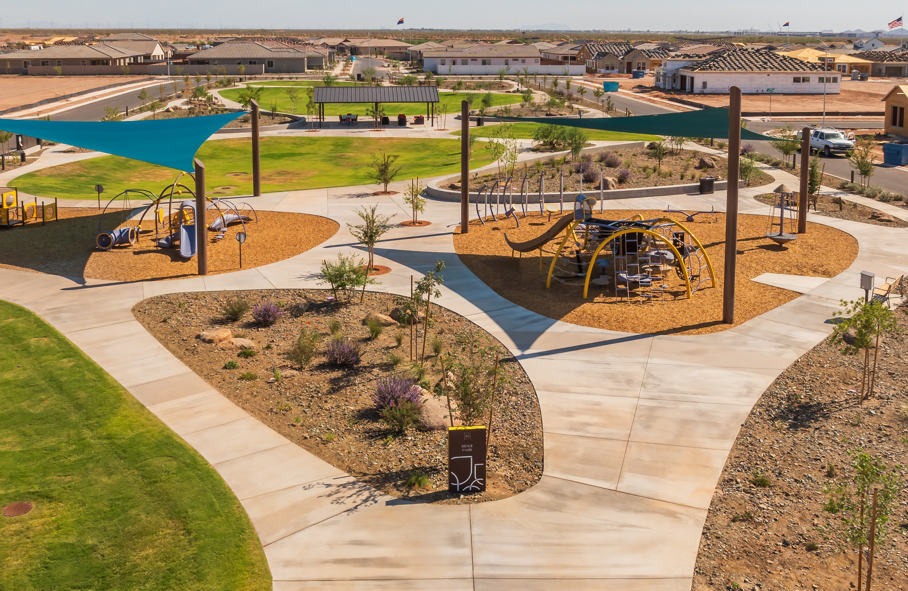 A vibrant and well-designed outdoor recreational area with various play structures, shaded seating areas, and lush landscaping, set against a backdrop of residential buildings.
