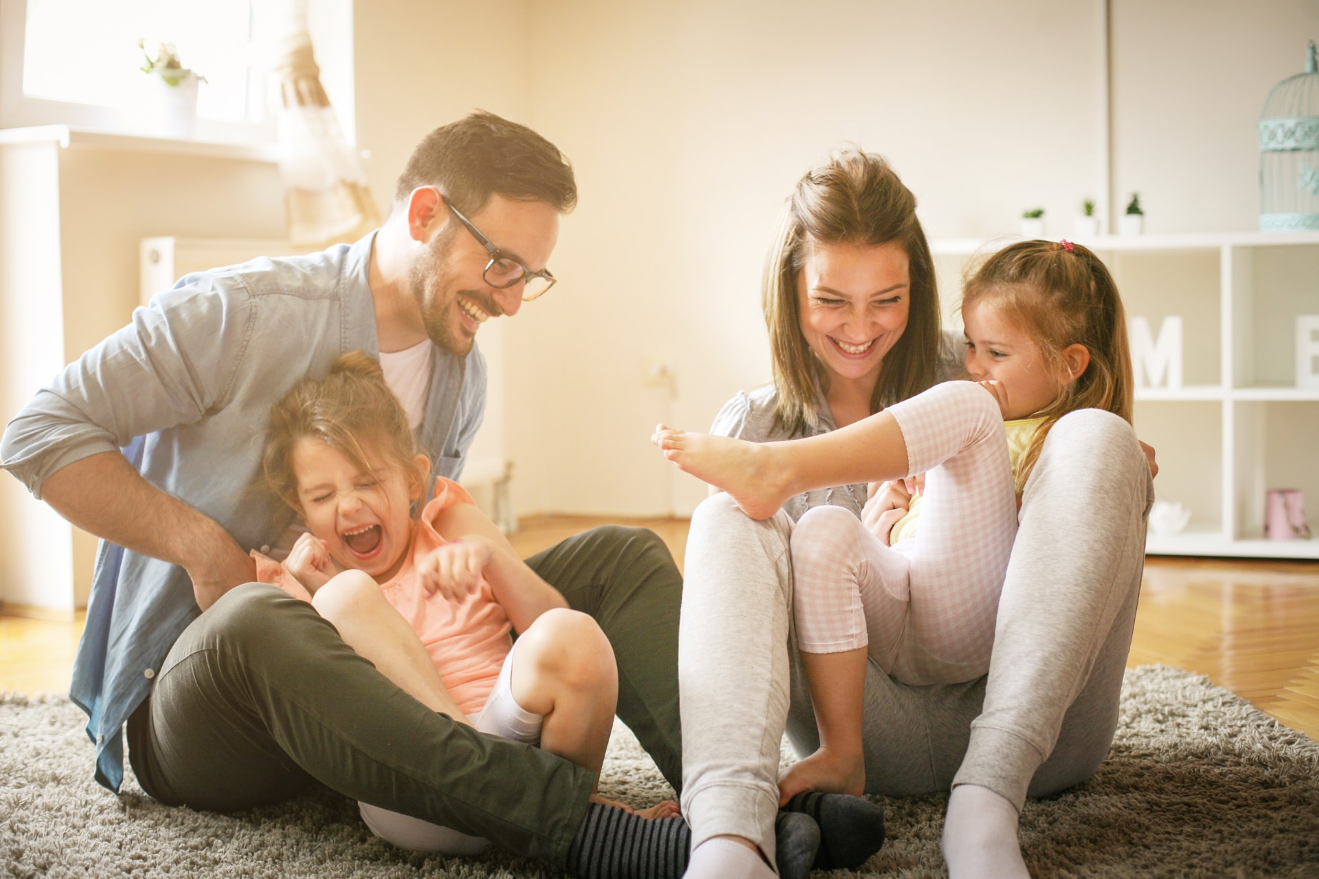 A family of four, including a mother, father, and two young daughters, are sitting together on the floor of a cozy, well-lit room, engaging in a playful and affectionate interaction.