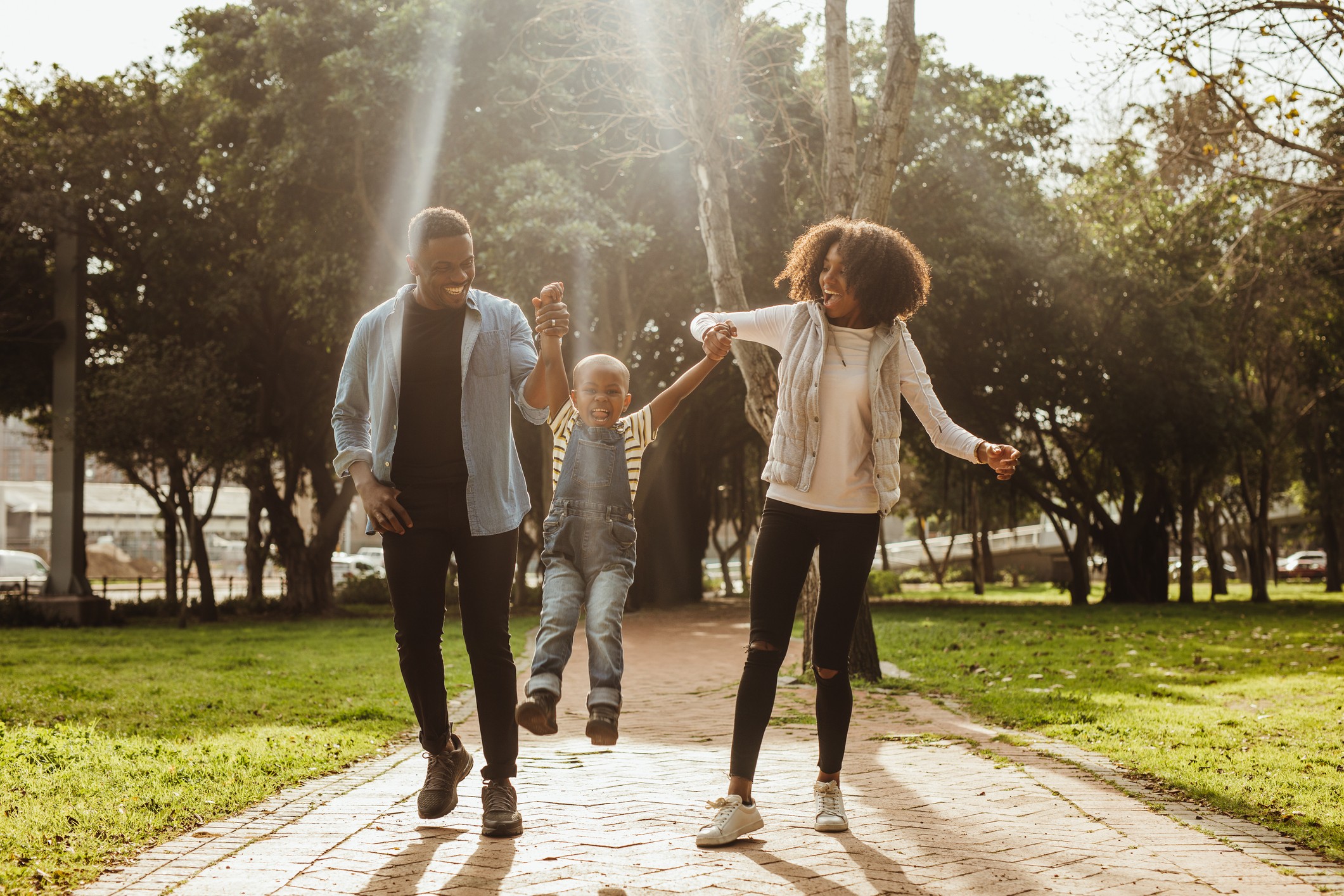 A family of four walks together on a paved path in a lush, green park, with sunlight filtering through the trees.
