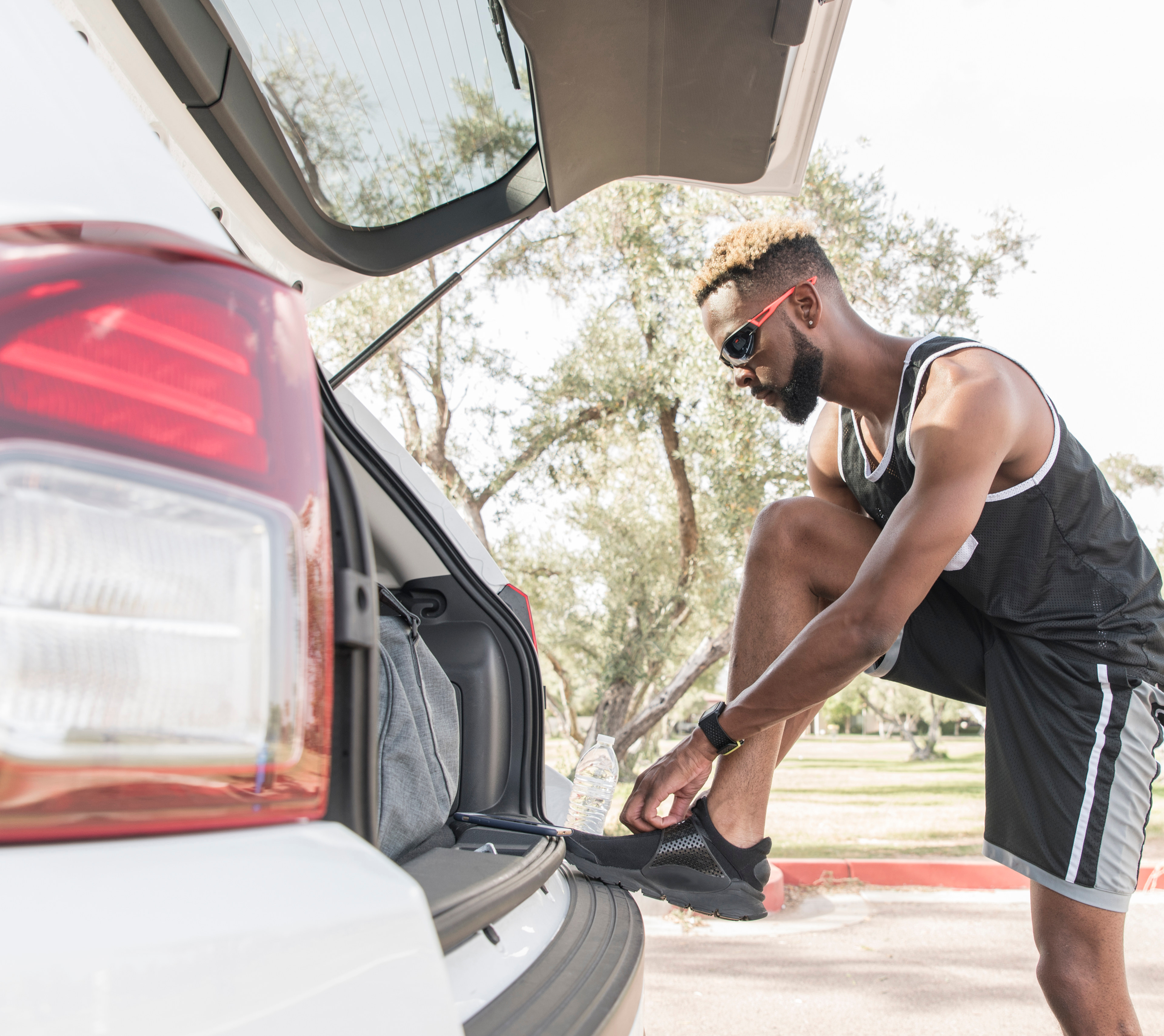A man in athletic clothing is leaning into the open trunk of a red car, surrounded by a grassy outdoor setting.