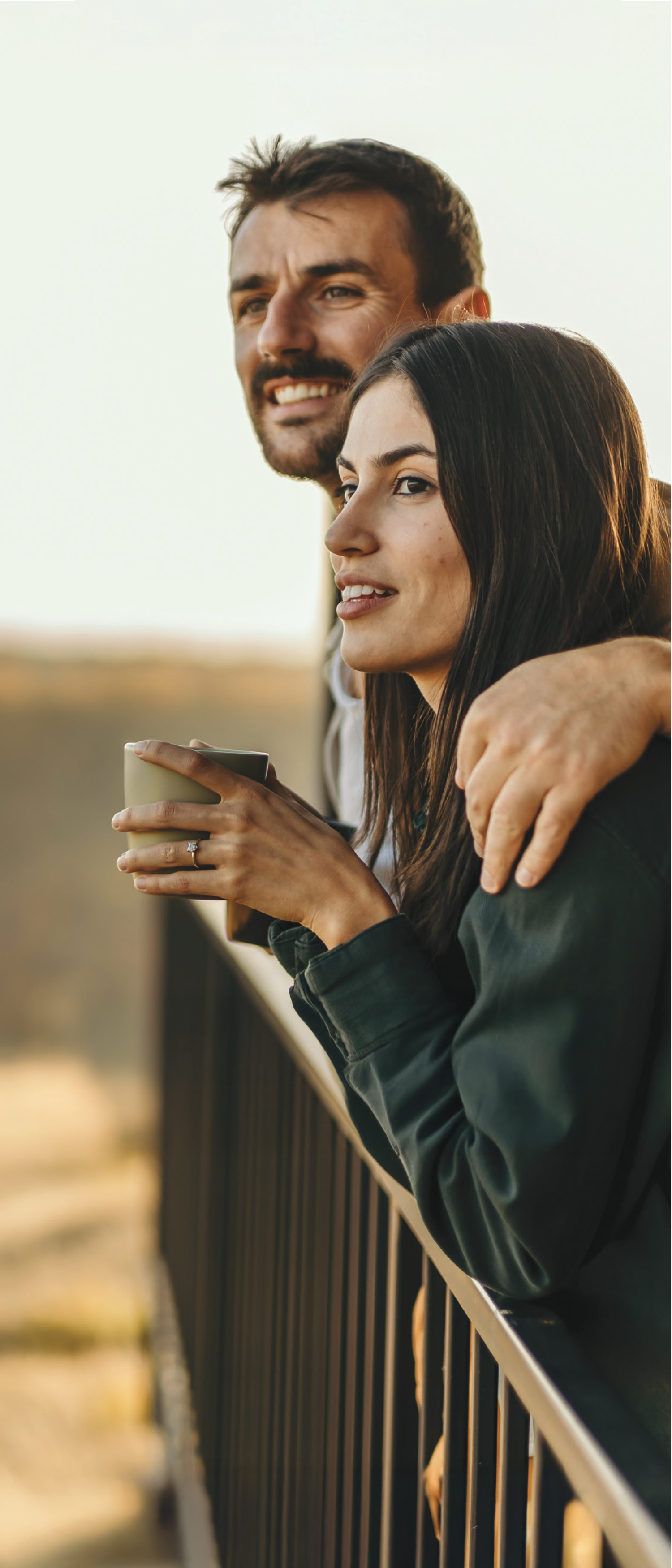 A smiling couple embracing and enjoying the view from a balcony or terrace, with a scenic landscape in the background.