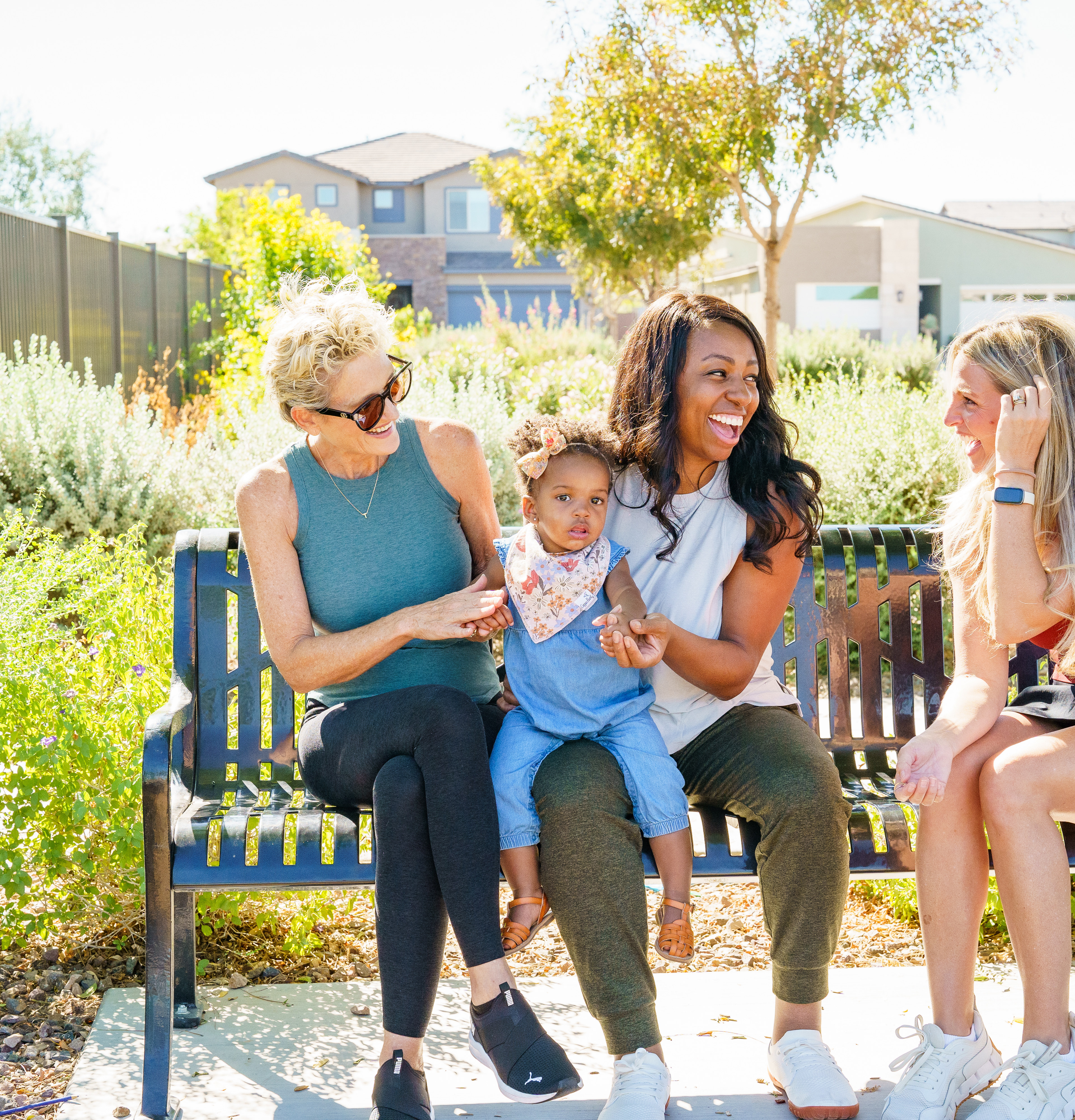 Three women sitting on a bench in a lush, green outdoor setting, with houses visible in the background.