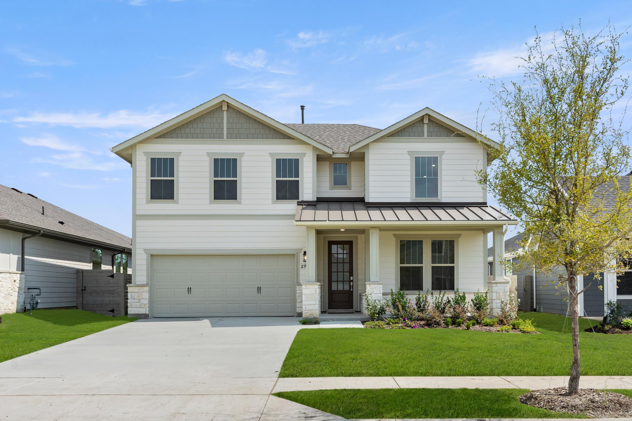 A two-story house with a gable roof, white siding, and a covered porch stands in a well-manicured yard with a grassy lawn and a young tree in the foreground.