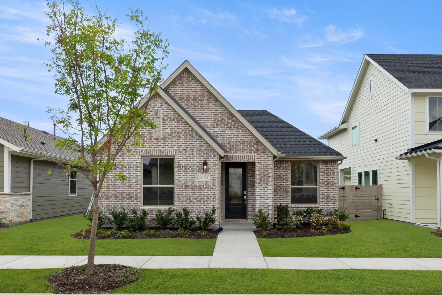 A newly constructed brick house with a gabled roof and a well-manicured lawn in the foreground, set against a backdrop of blue sky and lush greenery.
