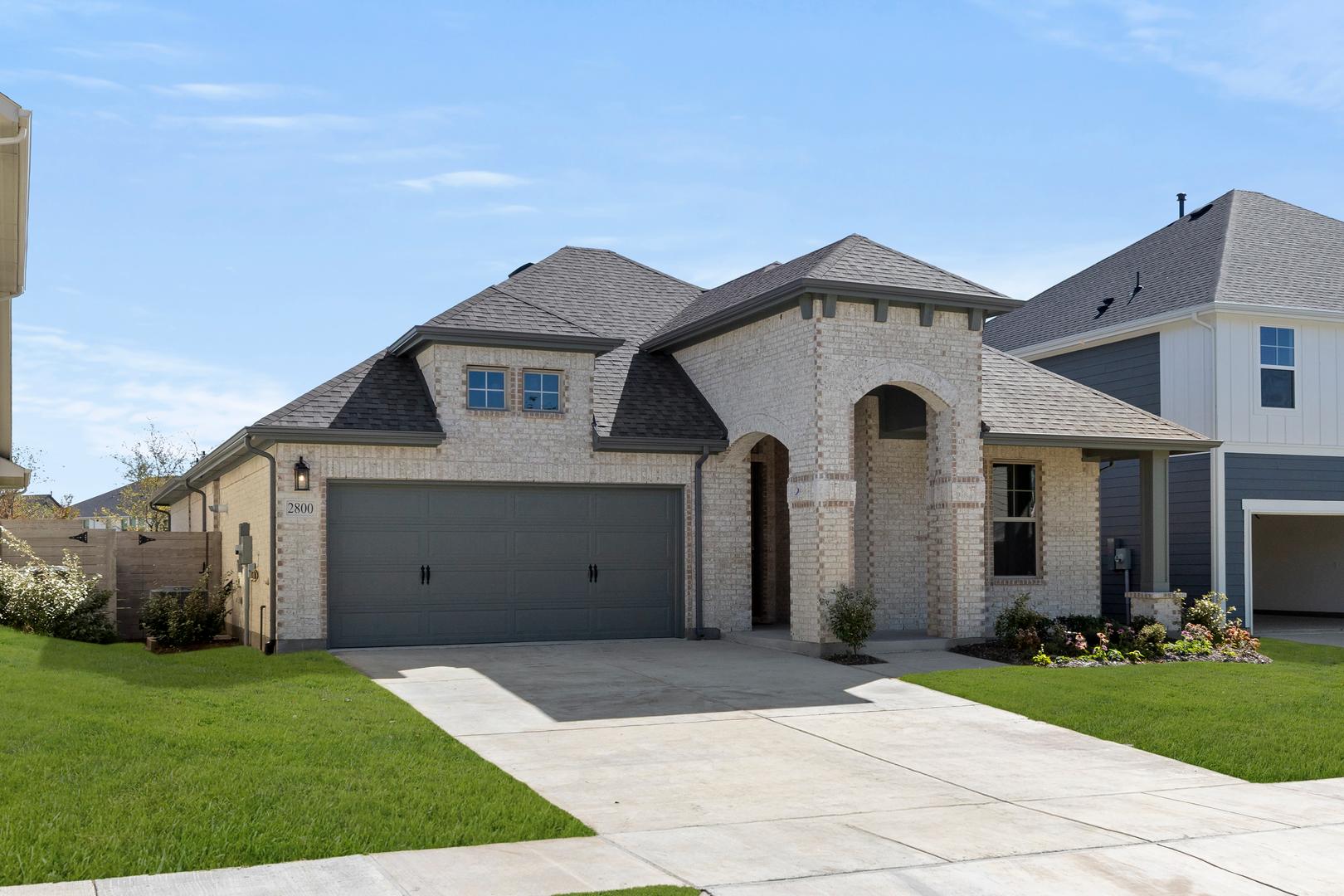 A large, two-story brick house with a prominent garage door and a well-manicured lawn in the foreground, set against a clear blue sky in the background.