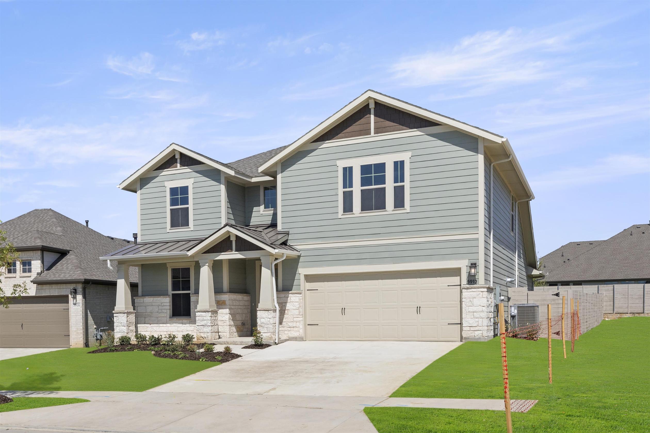 A two-story residential house with a gray exterior, a gabled roof, and a front porch, set against a clear blue sky with some clouds, surrounded by a grassy lawn and a wooden fence in the foreground.