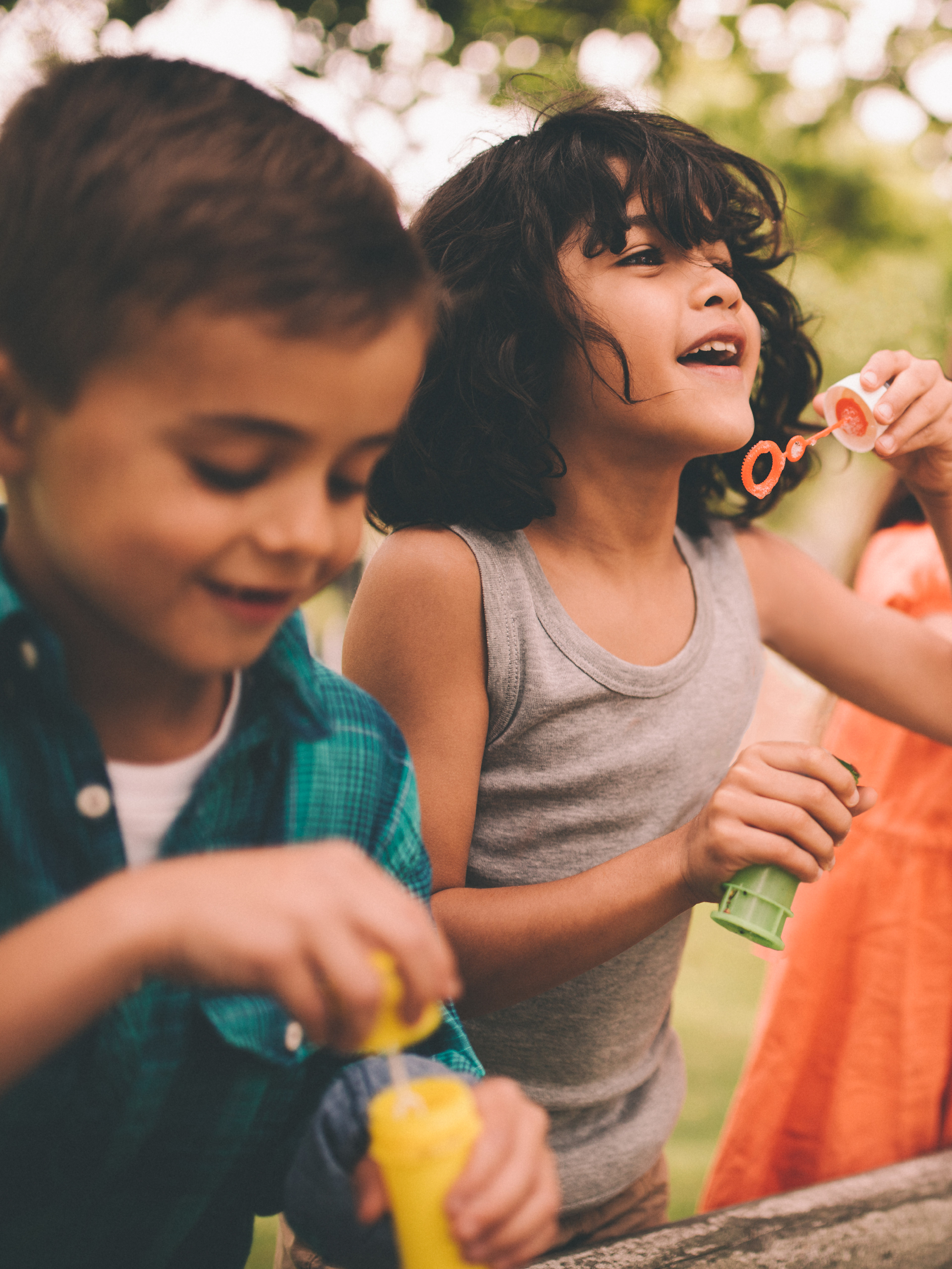 A group of young children playing with bubbles in a lush, outdoor setting with trees and greenery in the background.