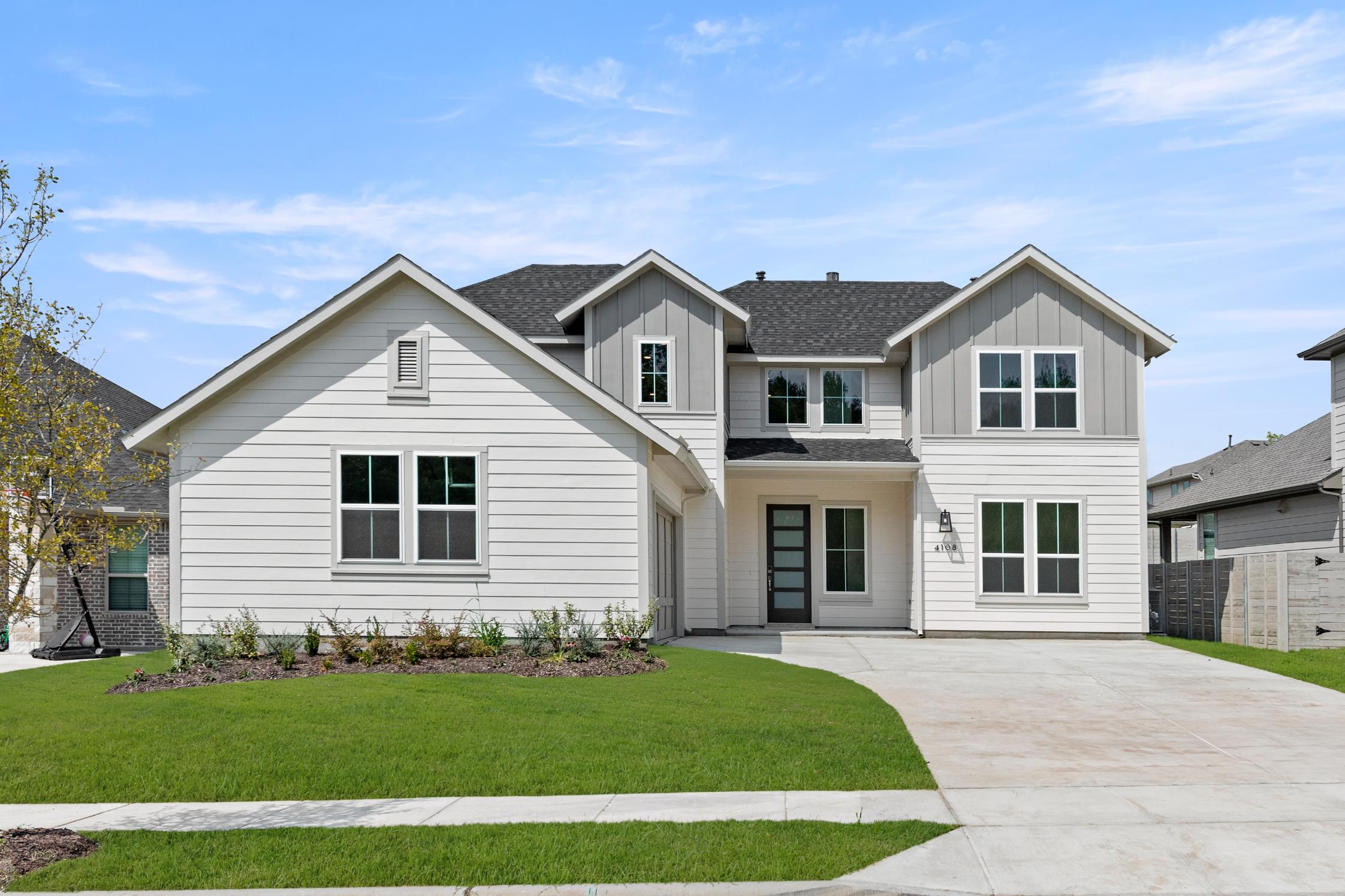 A two-story, gray-and-white house with a well-manicured lawn and a paved driveway, set against a clear blue sky with scattered clouds.