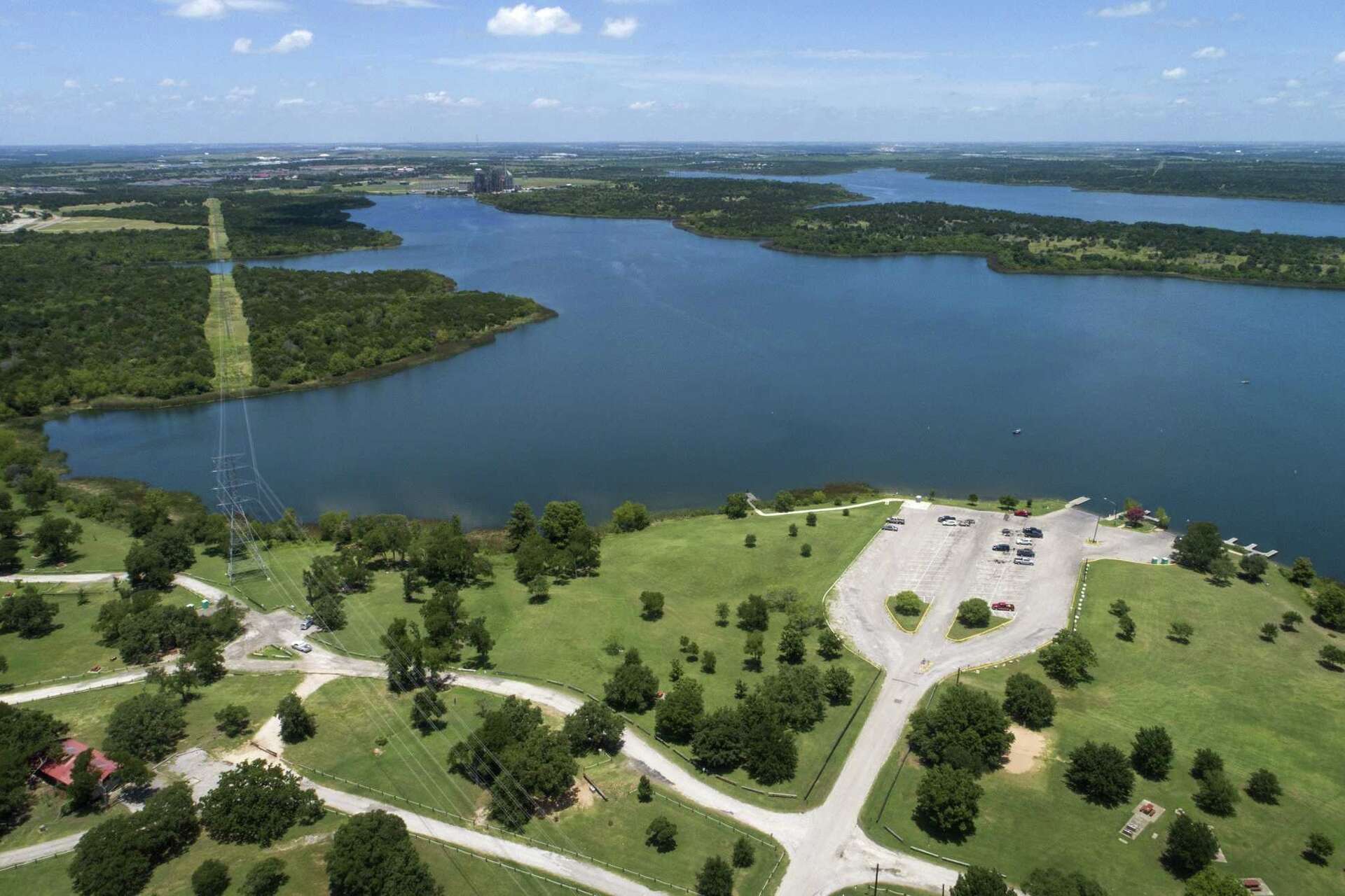 A scenic aerial view of a large lake surrounded by lush green forests, with a park-like area in the foreground featuring winding paths and a parking lot.