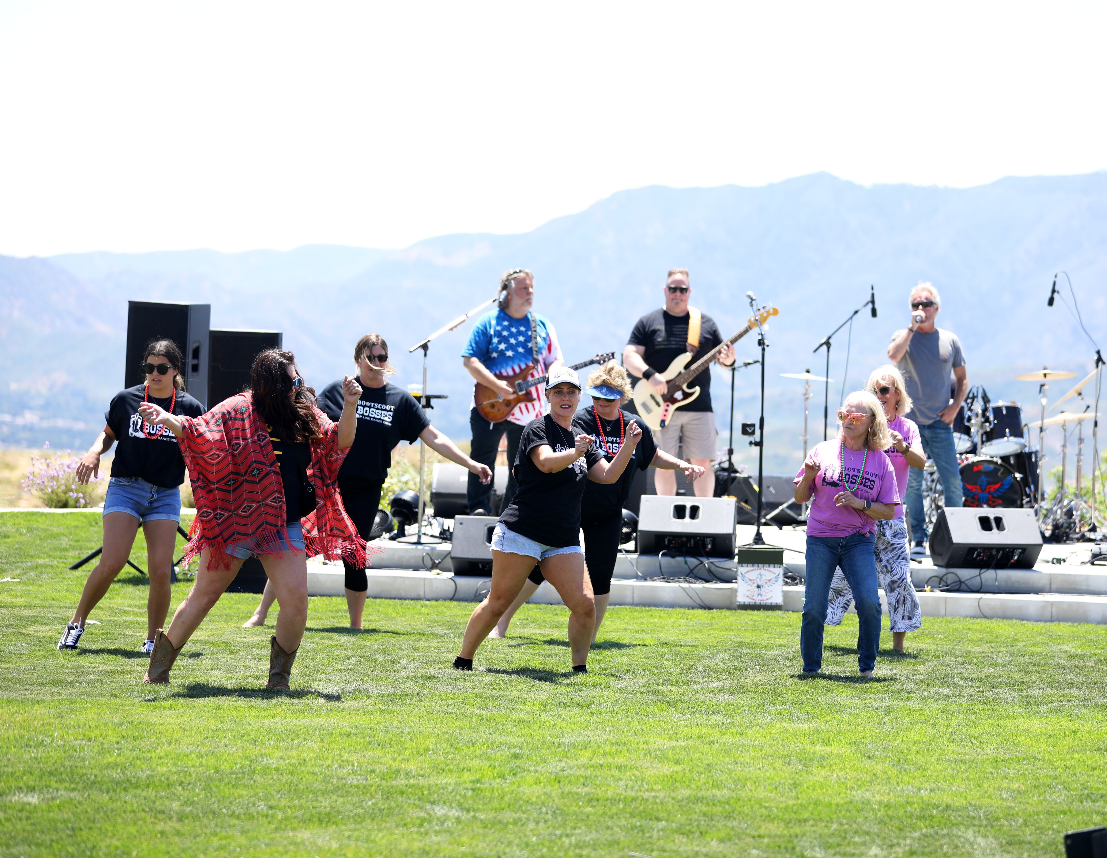 A group of people performing on a stage set up on a grassy field, with mountains visible in the background.