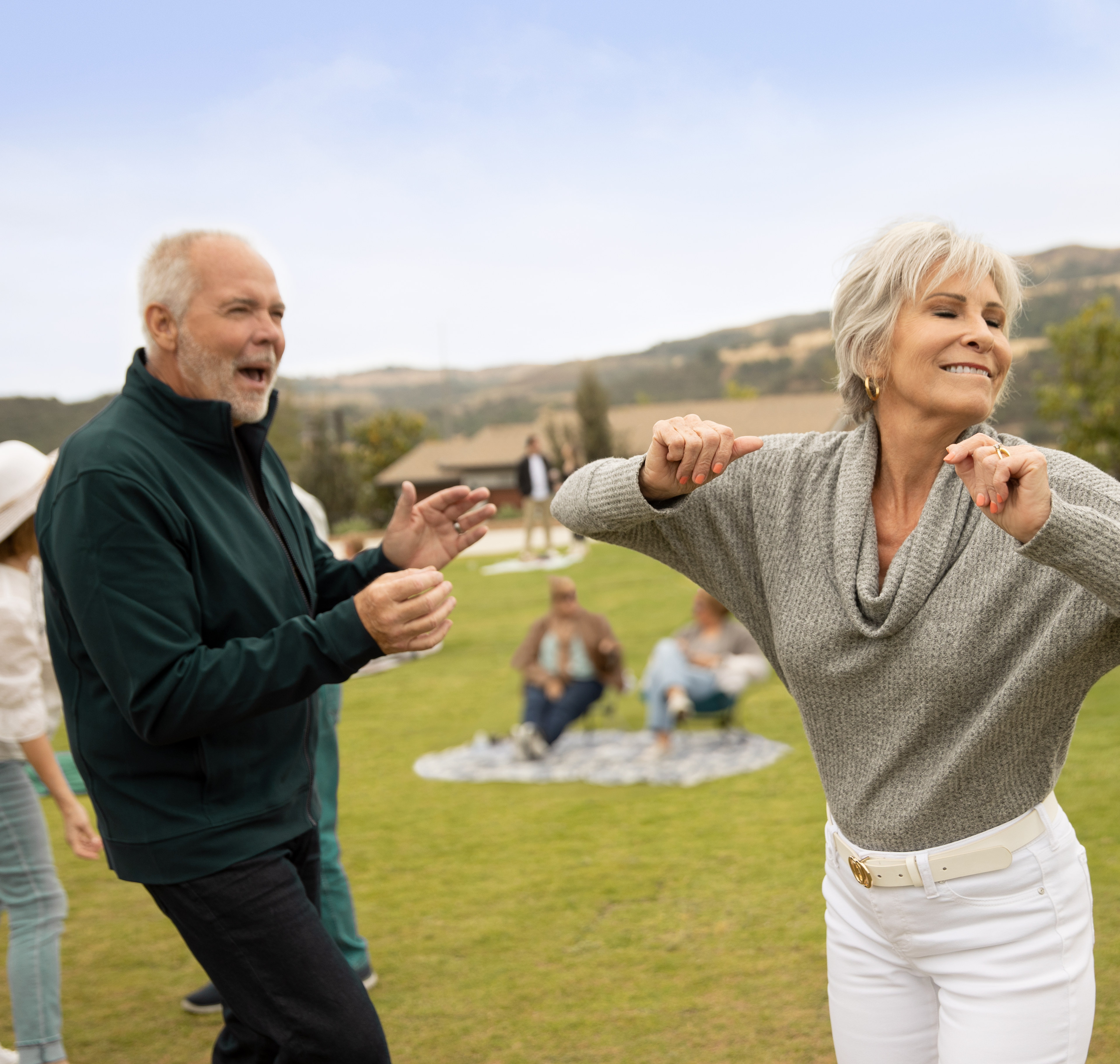 An elderly couple is dancing on a grassy field, with a group of people in the background enjoying the outdoors.
