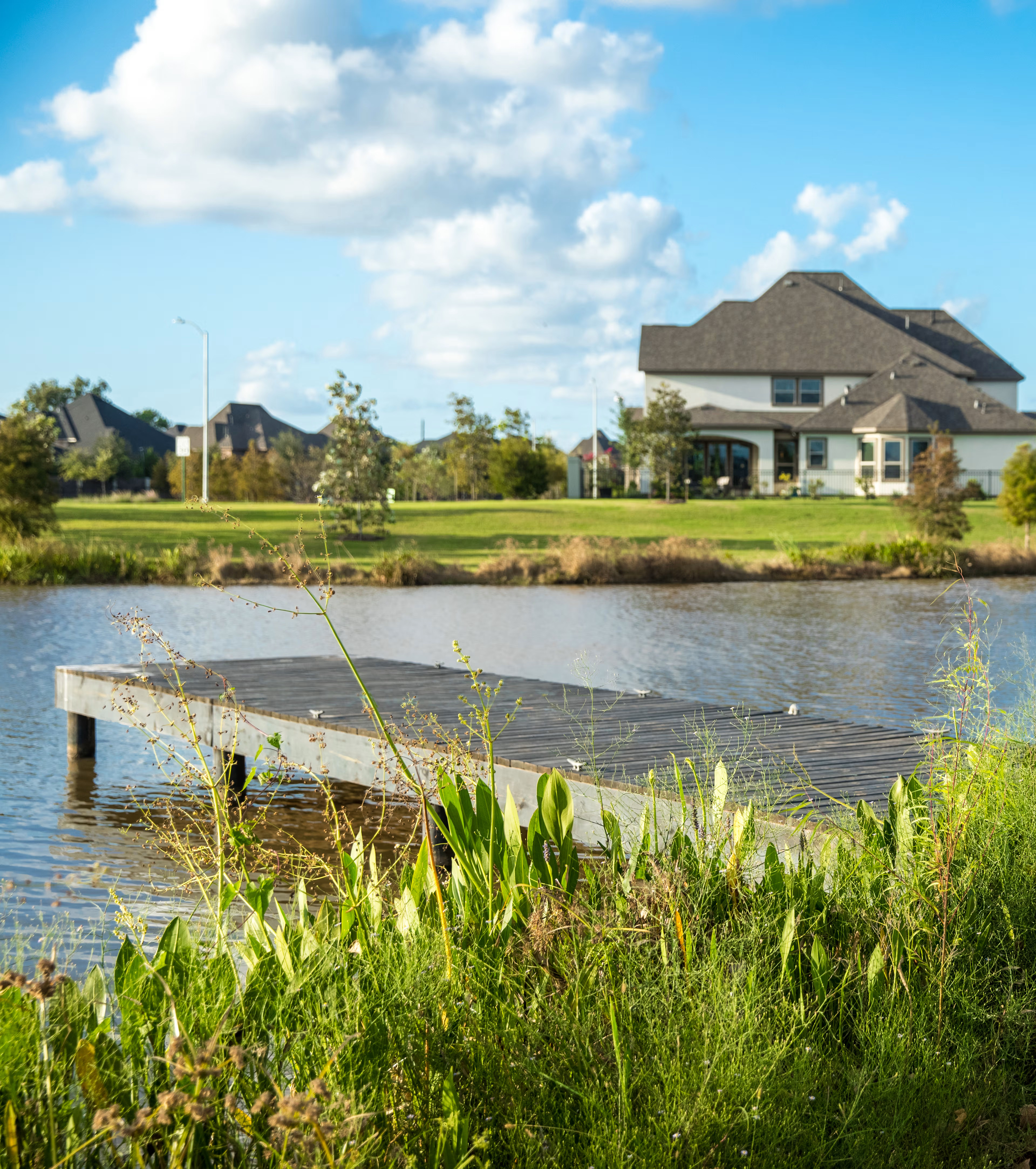 A picturesque suburban neighborhood with a large house overlooking a tranquil pond, surrounded by lush greenery and a wooden dock in the foreground.