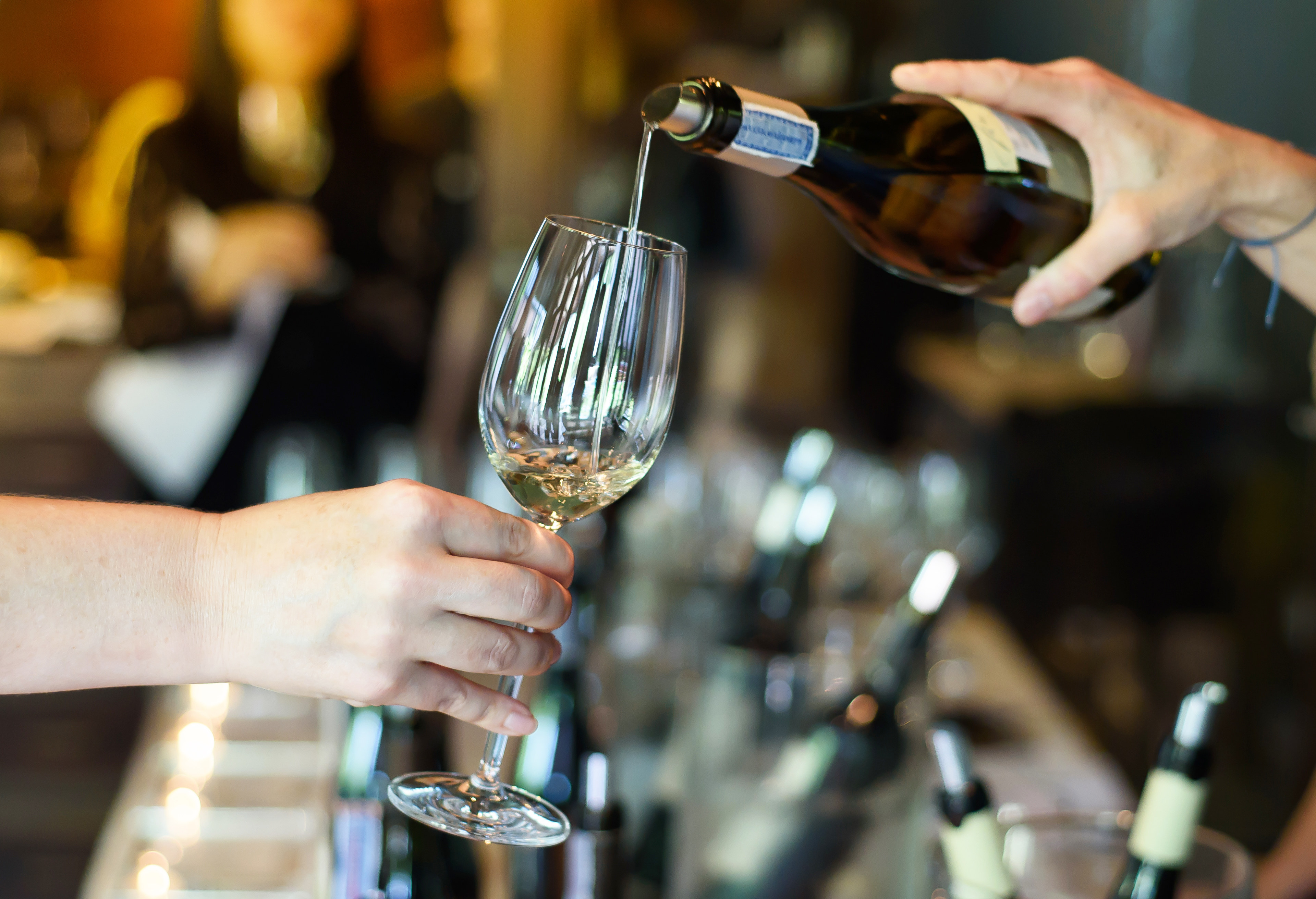 A person's hands are pouring wine into a crystal wine glass against a backdrop of wine bottles and glasses.
