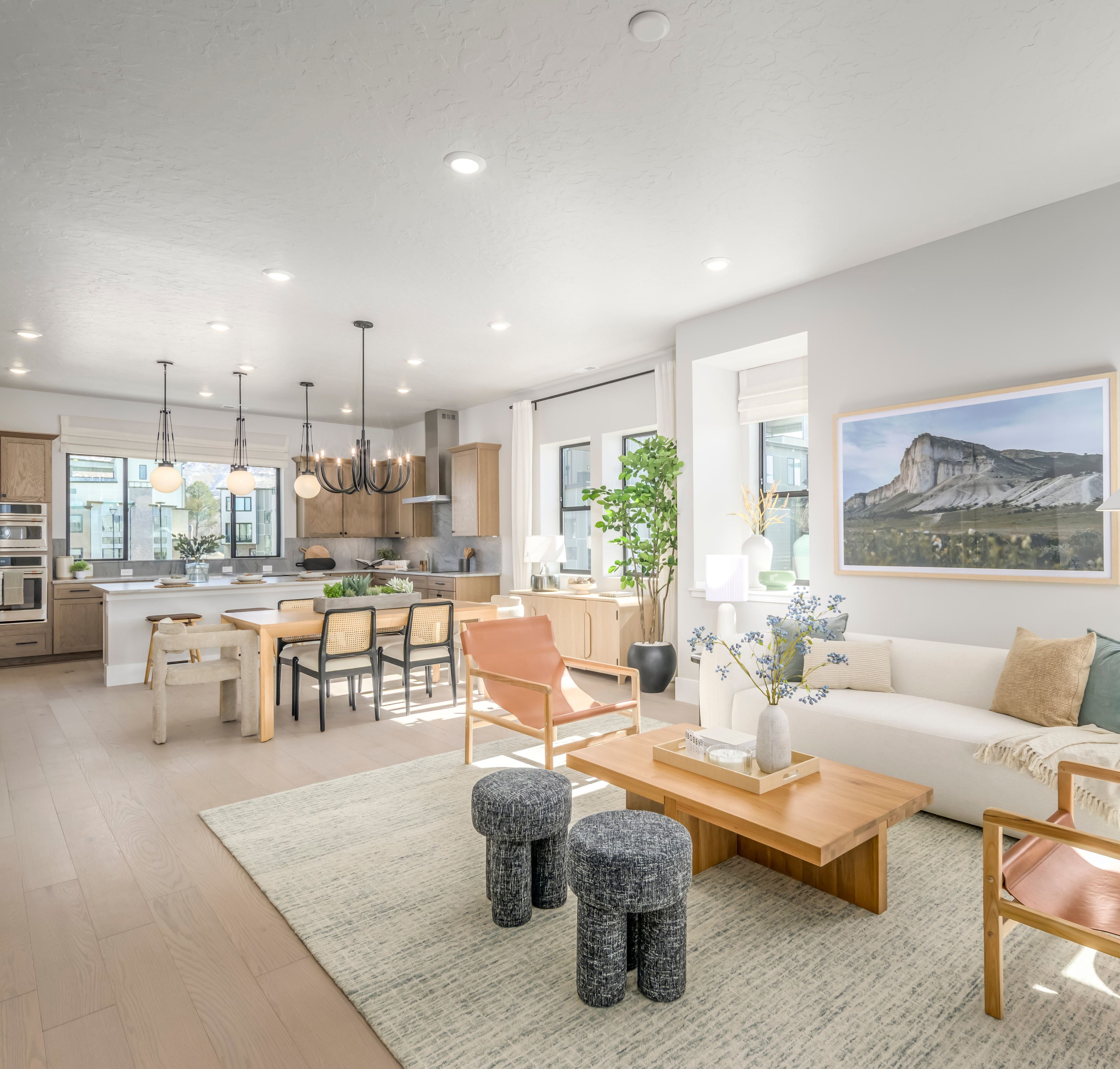 A modern and spacious open-concept living area with a kitchen, dining, and living room, featuring light-colored furniture, large windows, and a scenic landscape artwork on the wall.