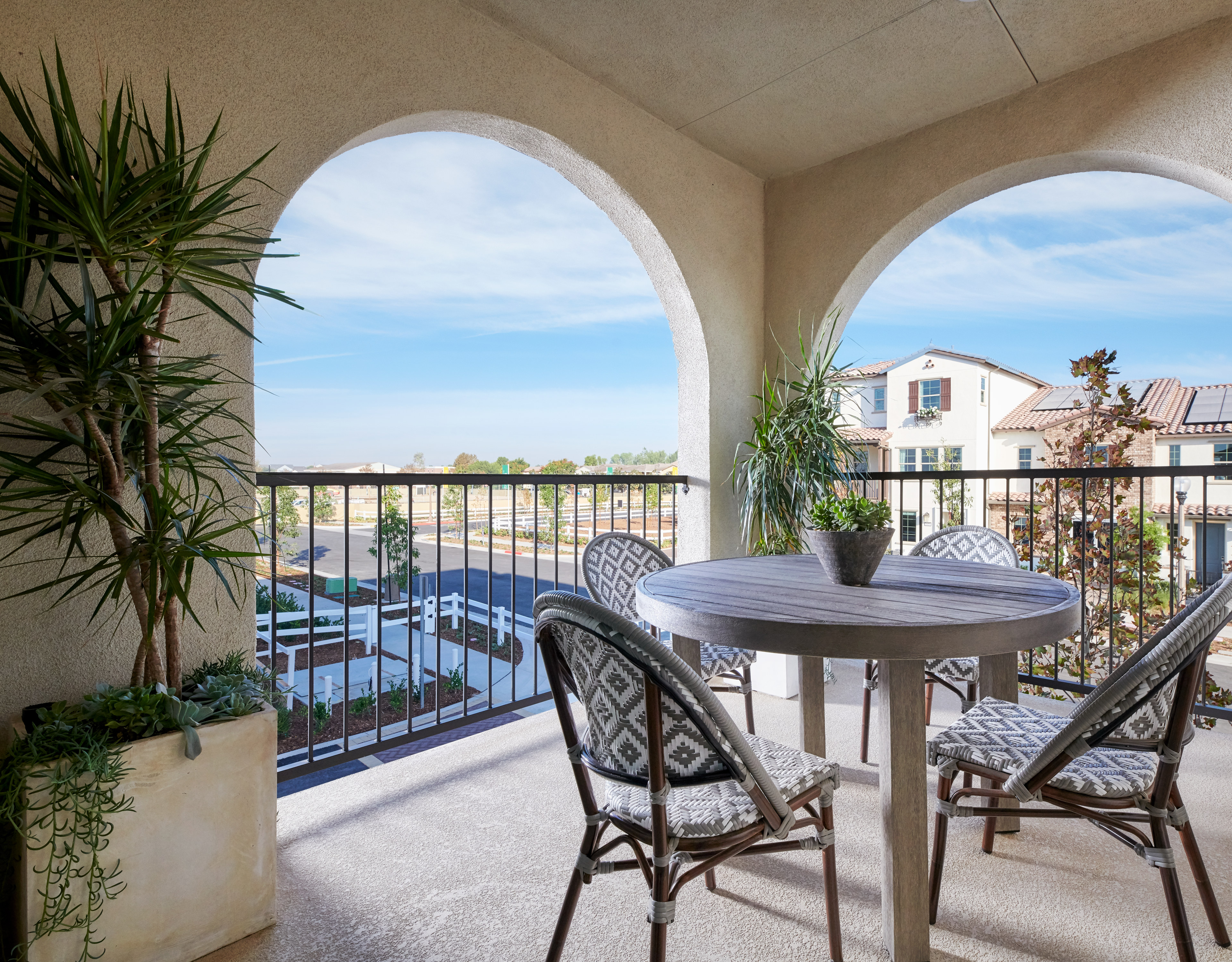 A cozy balcony with arched windows overlooking a scenic outdoor area, featuring a round table and chairs surrounded by potted plants.
