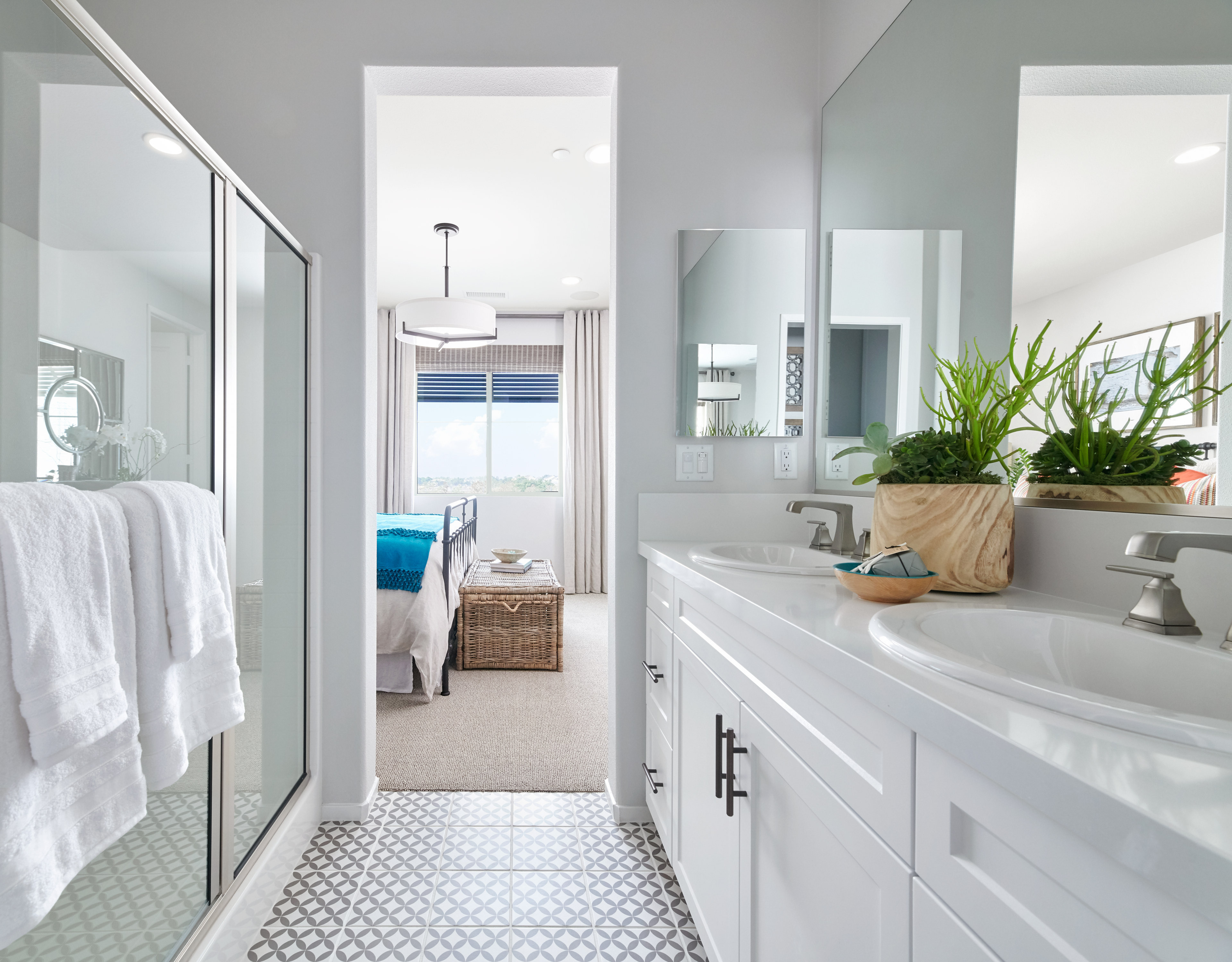A bright and airy bathroom with white cabinets, a large mirror, and a potted plant, leading to an open doorway in the background.