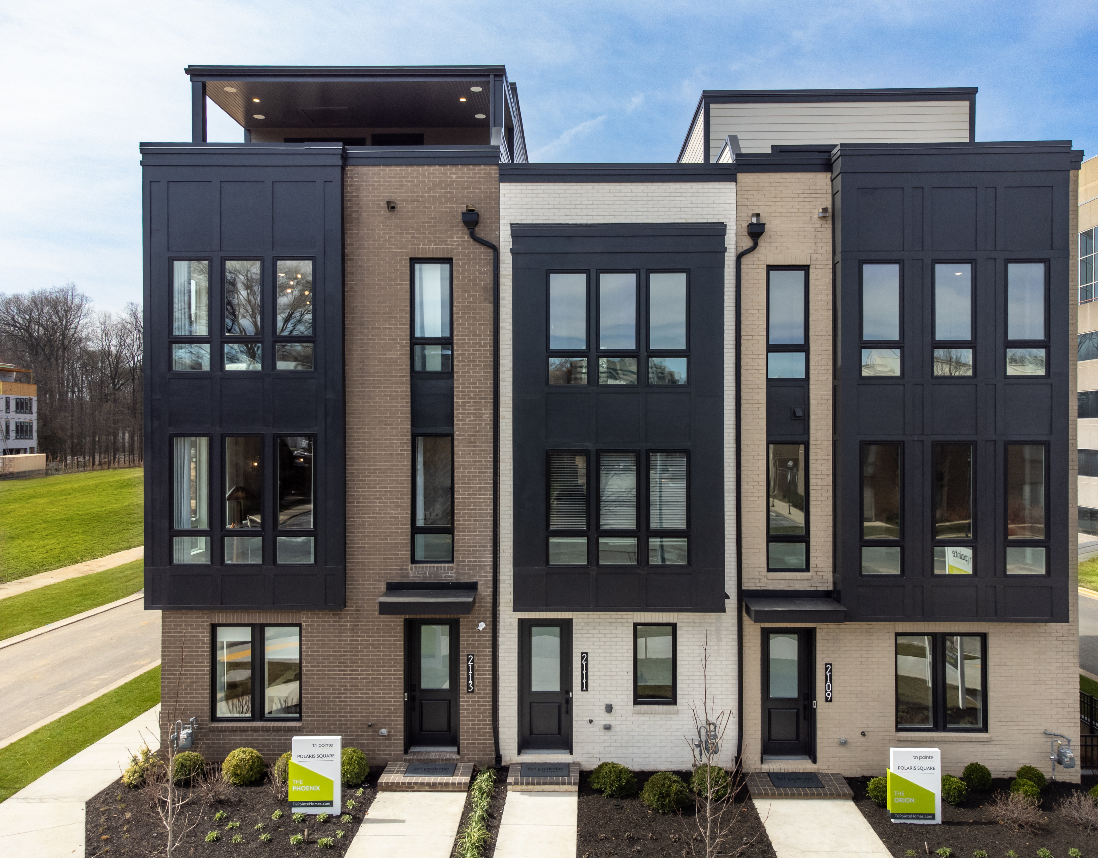 A modern, multi-story townhouse complex with a sleek, dark exterior and large windows, set against a backdrop of a grassy lawn and clear blue sky.