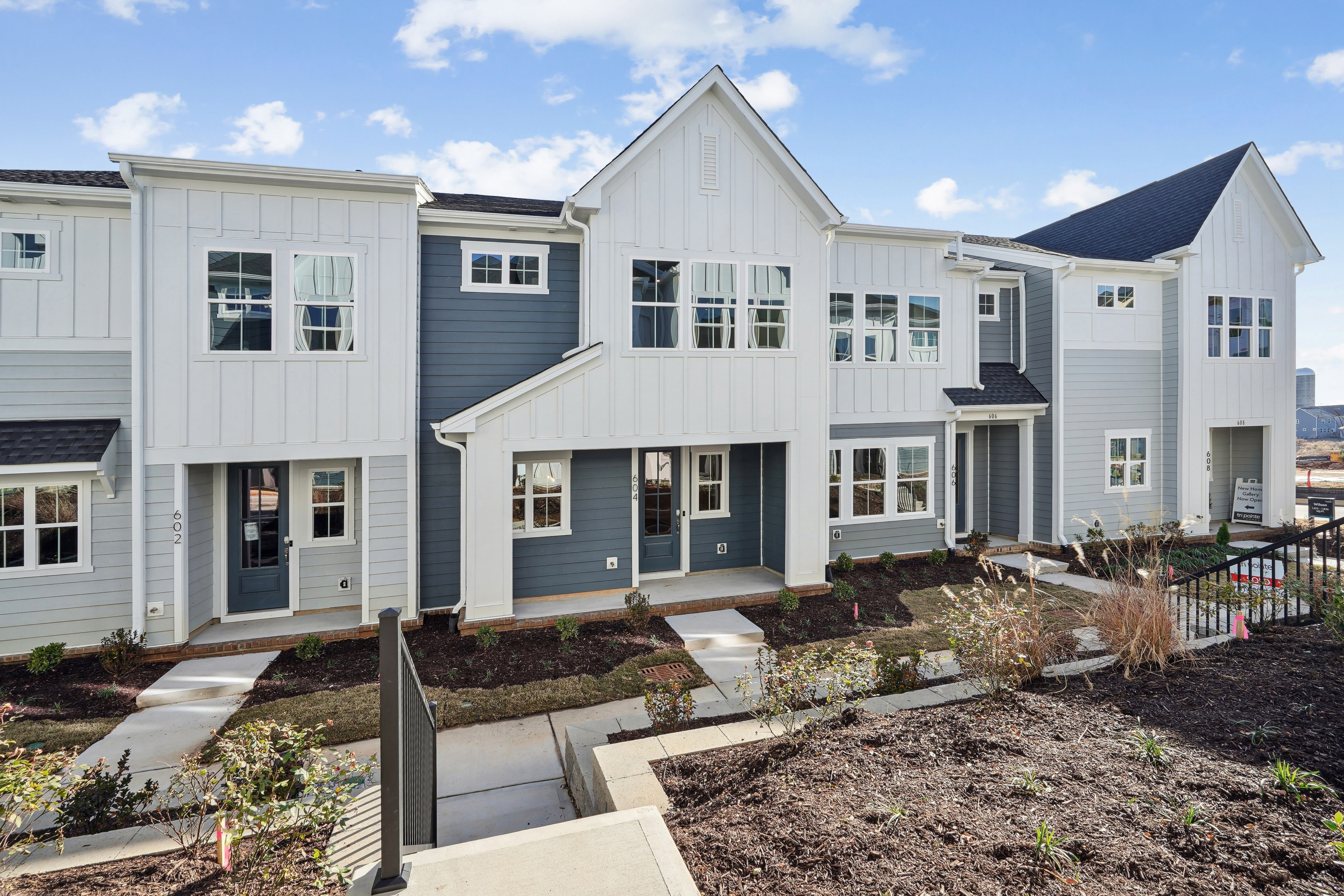 A row of modern, two-story townhouses with white siding, gray accents, and gabled roofs, set against a backdrop of a blue sky with scattered clouds.