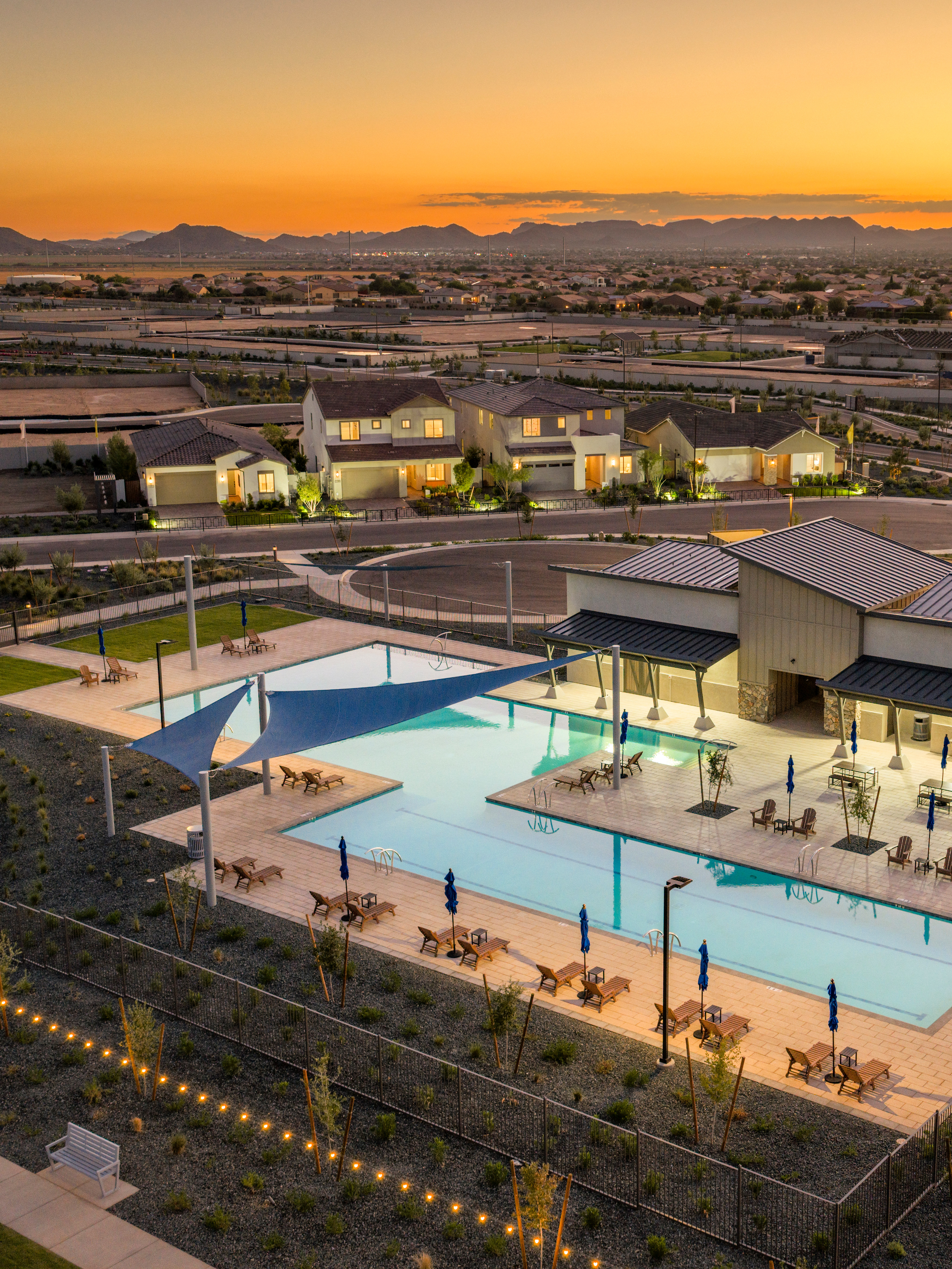 A residential community with a swimming pool and surrounding buildings, set against a backdrop of mountains and a vibrant sunset sky.