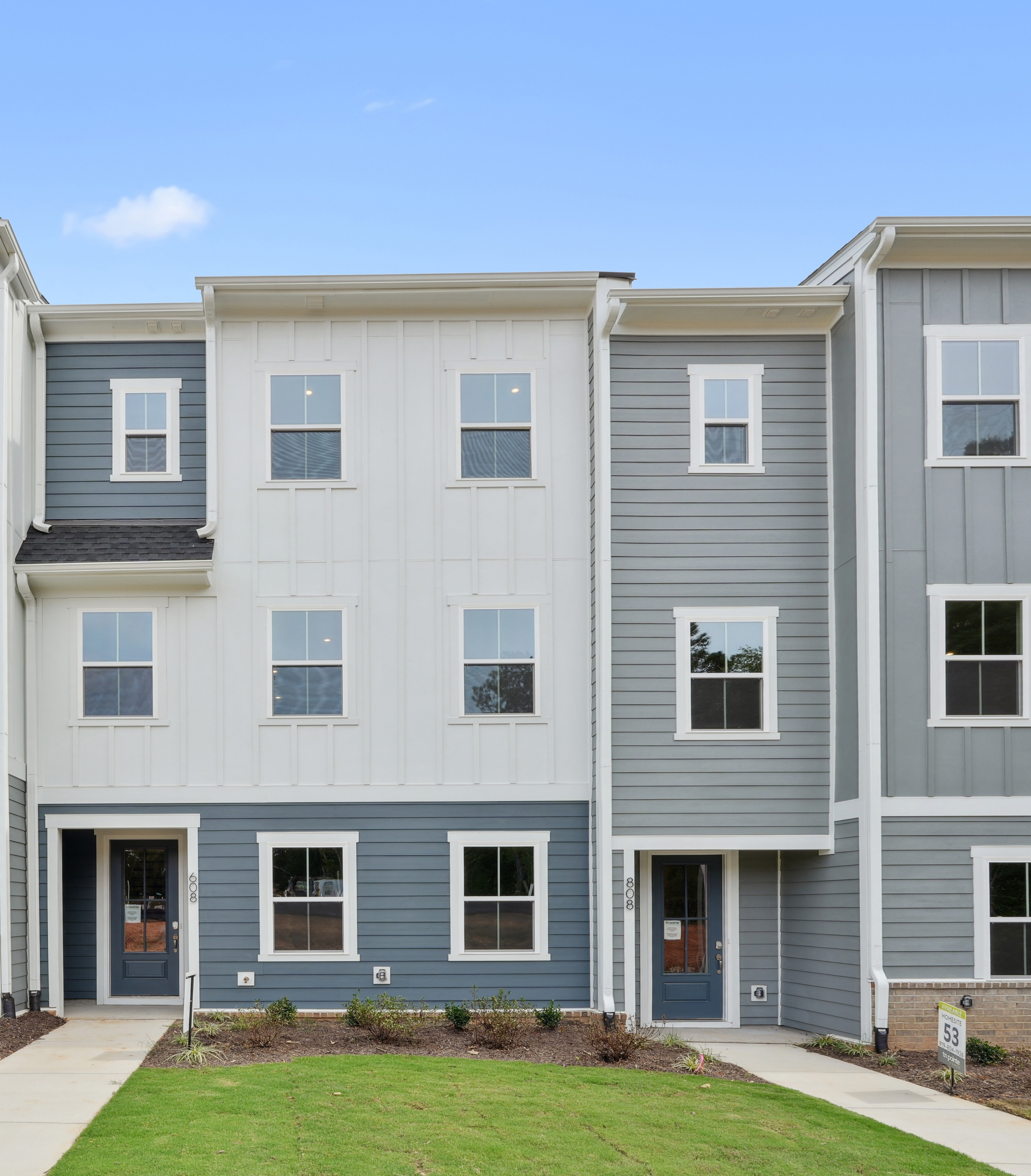 A multi-story townhouse complex with gray siding, white trim, and a grassy lawn in the foreground.