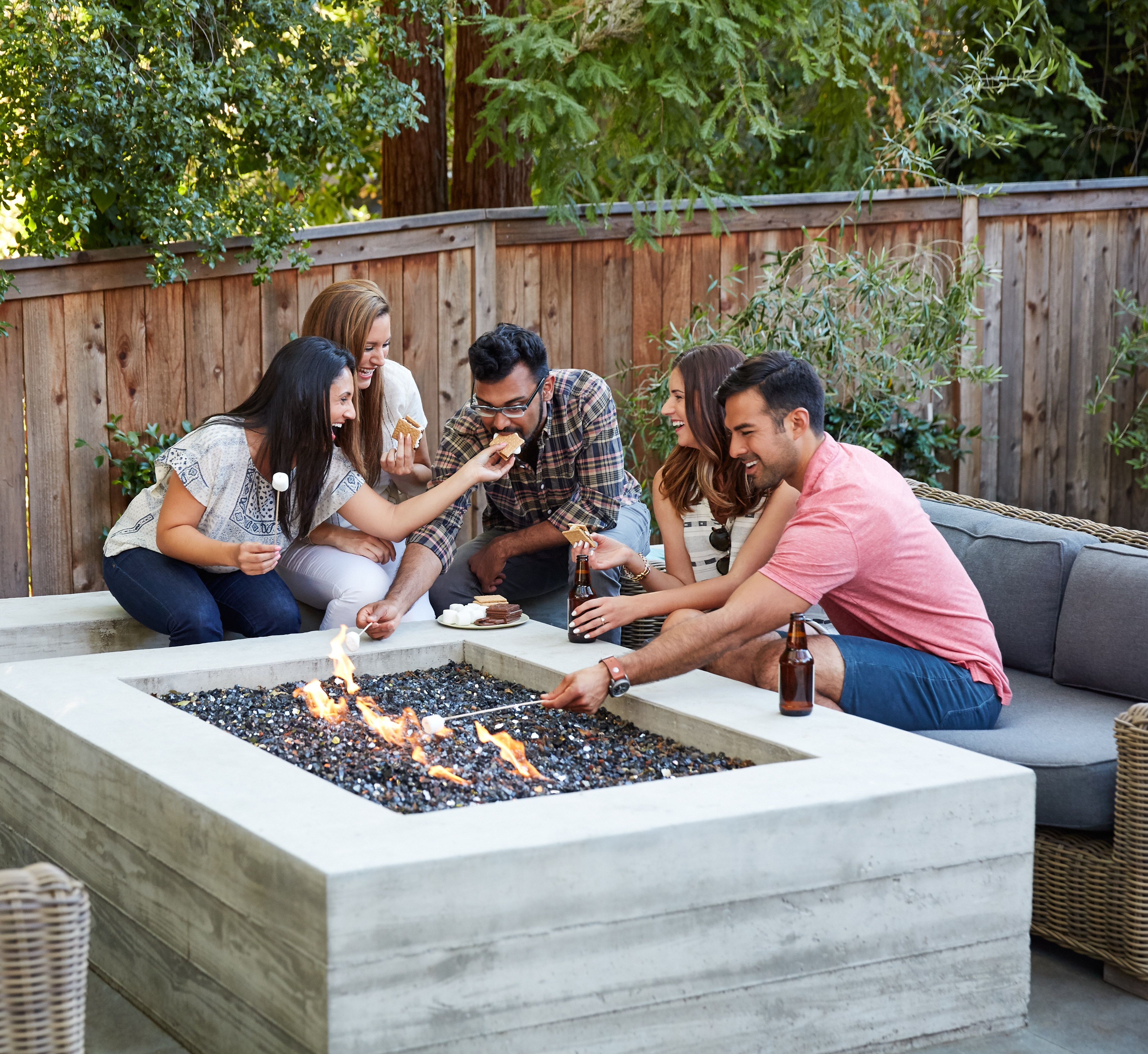 A group of friends gathered around a concrete fire pit, enjoying each other's company in a cozy outdoor setting surrounded by lush greenery and a wooden fence.