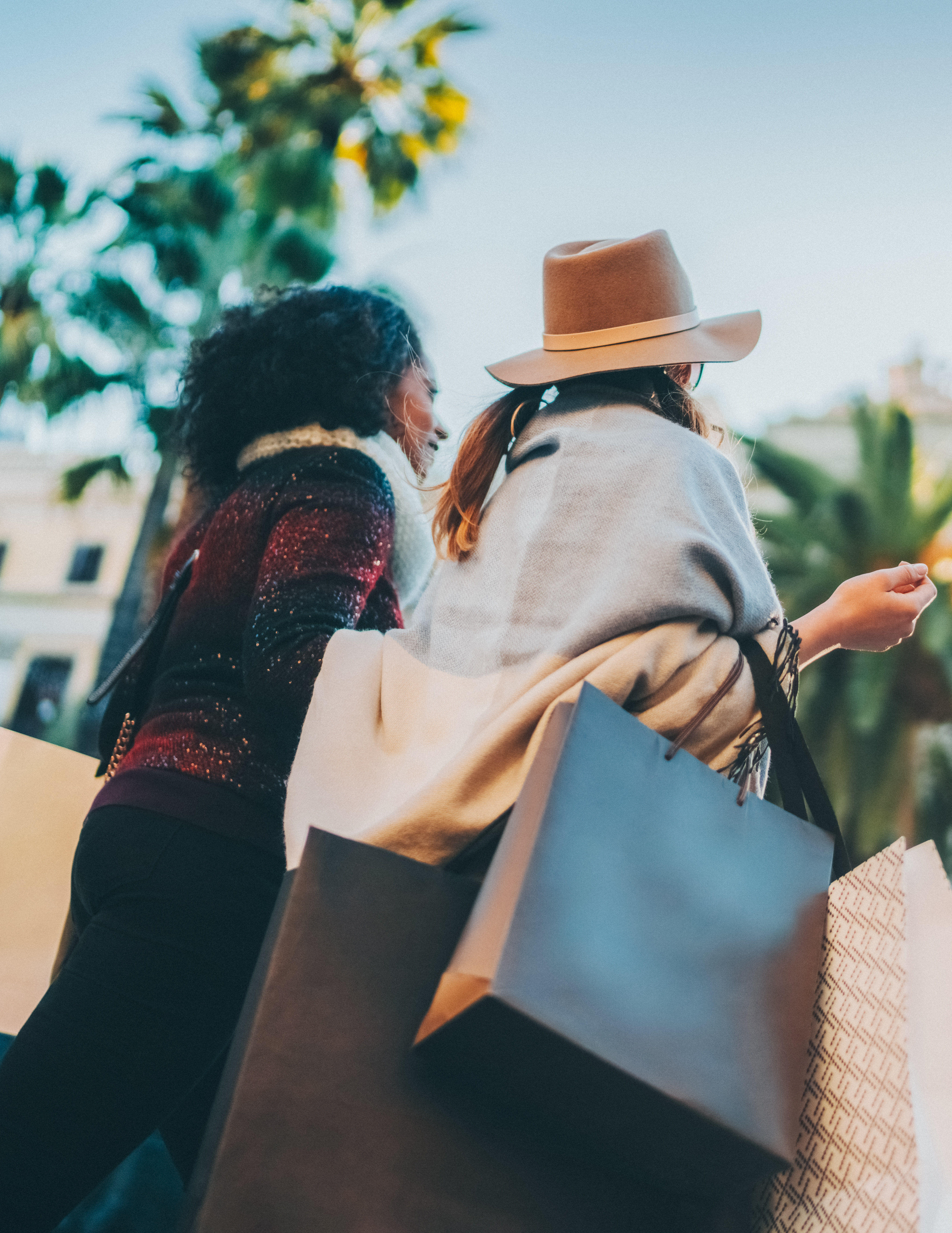 Two people, a man and a woman, are walking together while carrying shopping bags in a sunny, palm tree-lined street.