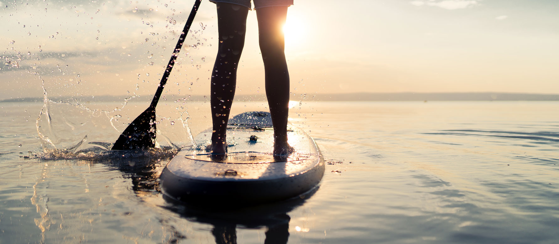 A person's legs and feet are visible in the foreground, standing on a paddleboard on a calm body of water, with a serene sunset in the background.