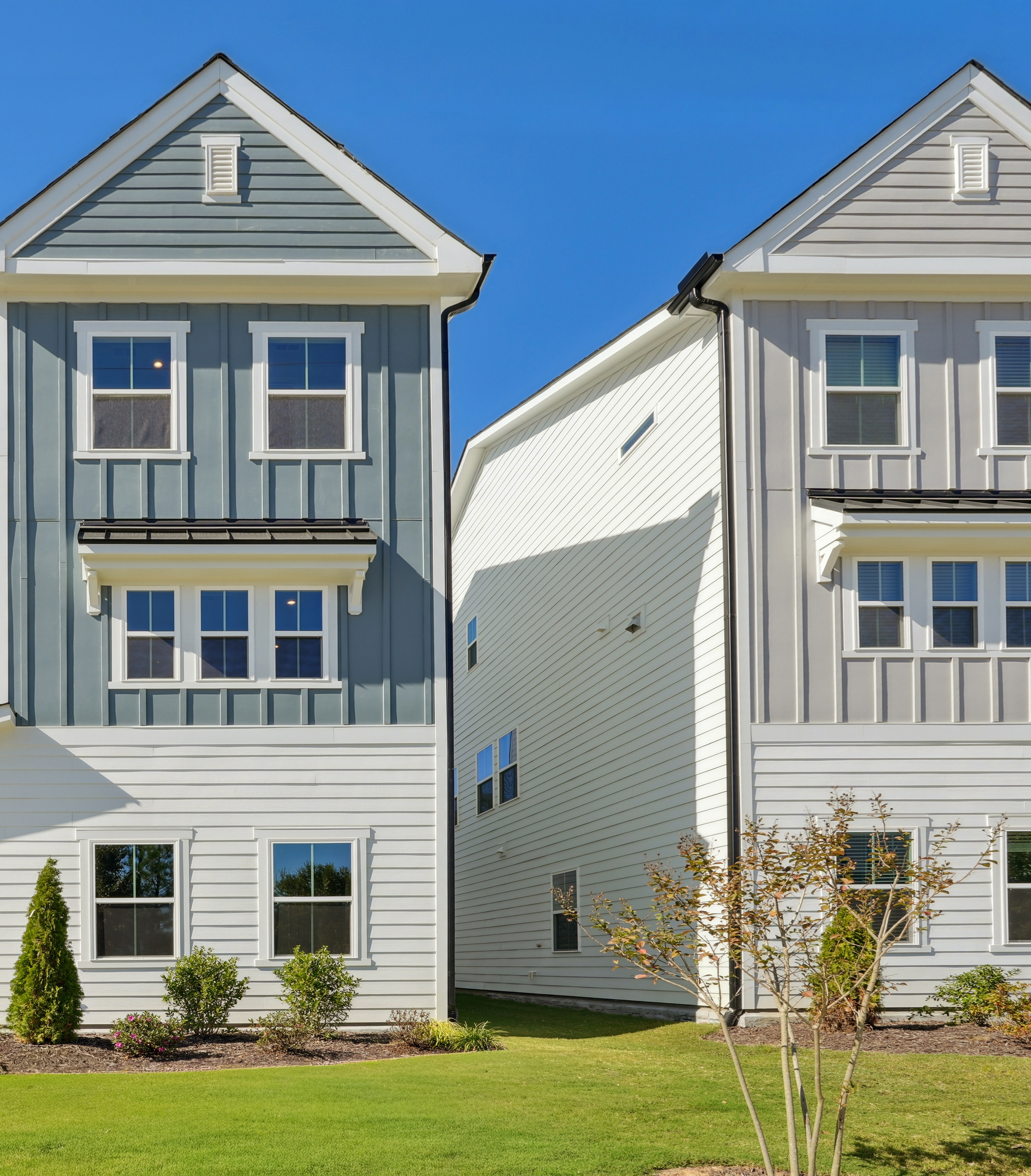 A row of modern, two-story townhouses with gray siding, white trim, and gabled roofs set against a clear blue sky, surrounded by lush greenery in the foreground.