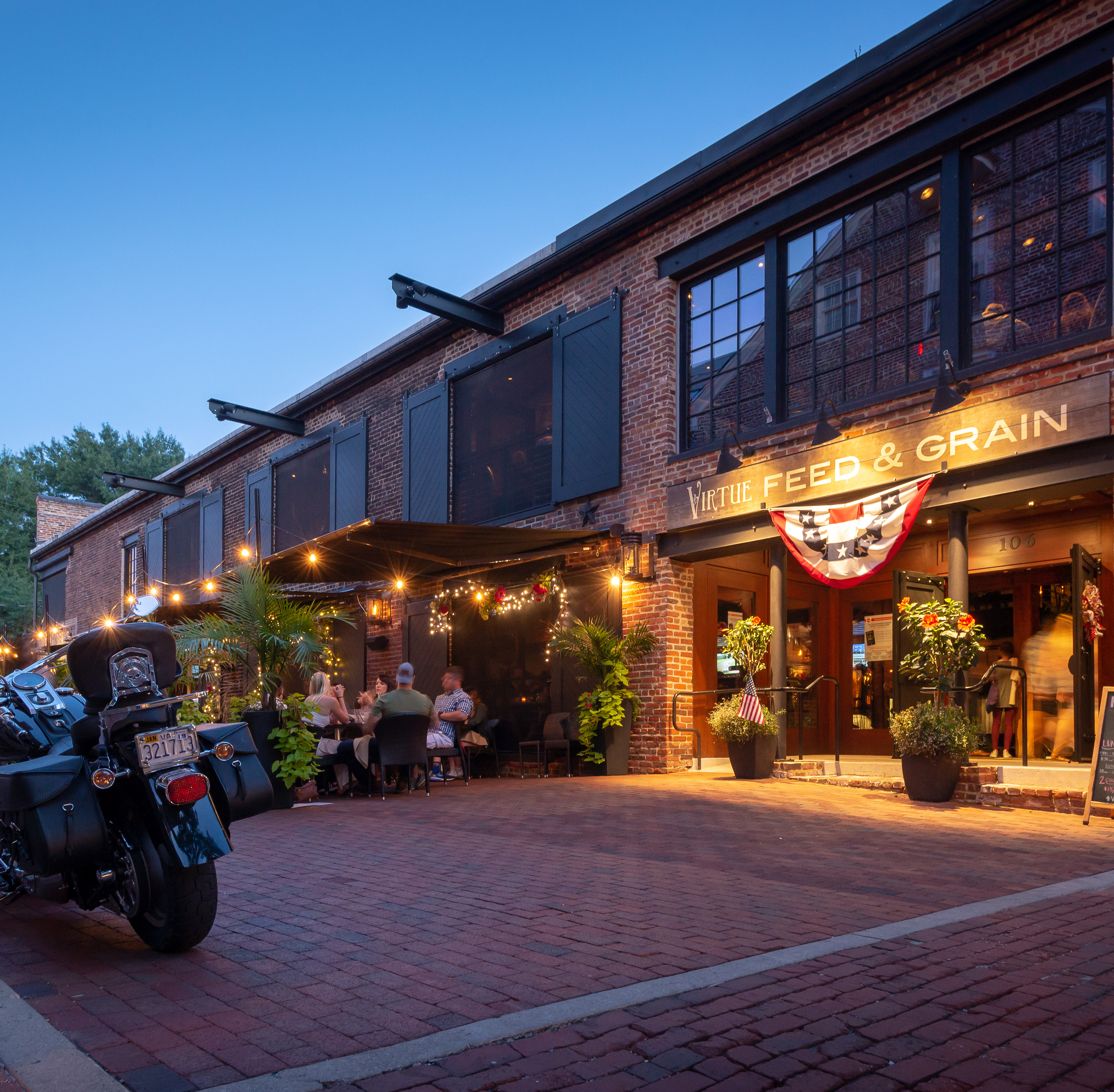 A lively street scene with a brick building housing a restaurant called "Seed & Grain" and several people walking along the sidewalk, with a motorcycle parked in the foreground.