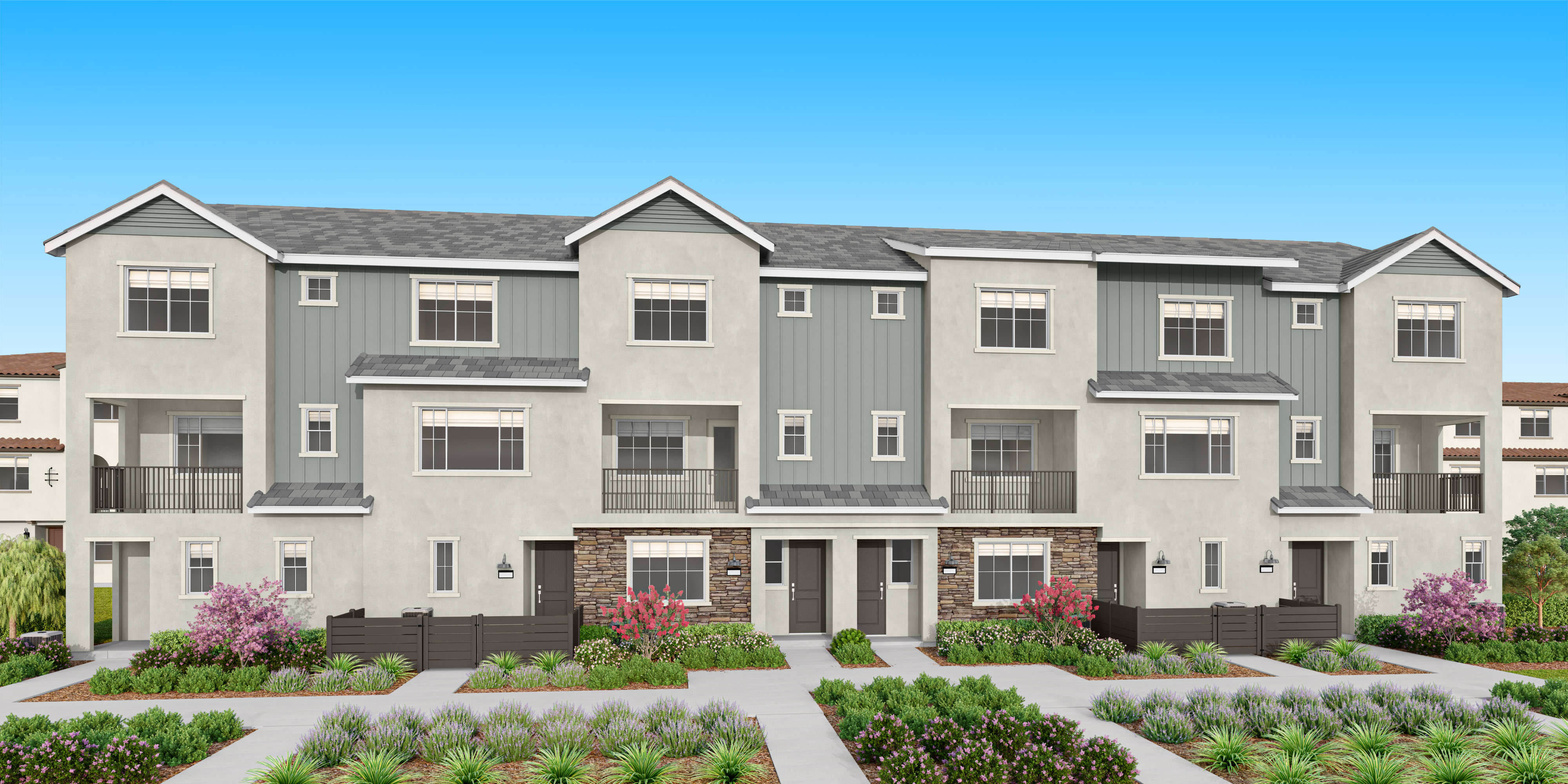 A multi-story townhouse complex with gray siding, red roofs, and landscaped yards in the foreground, set against a clear blue sky in the background.