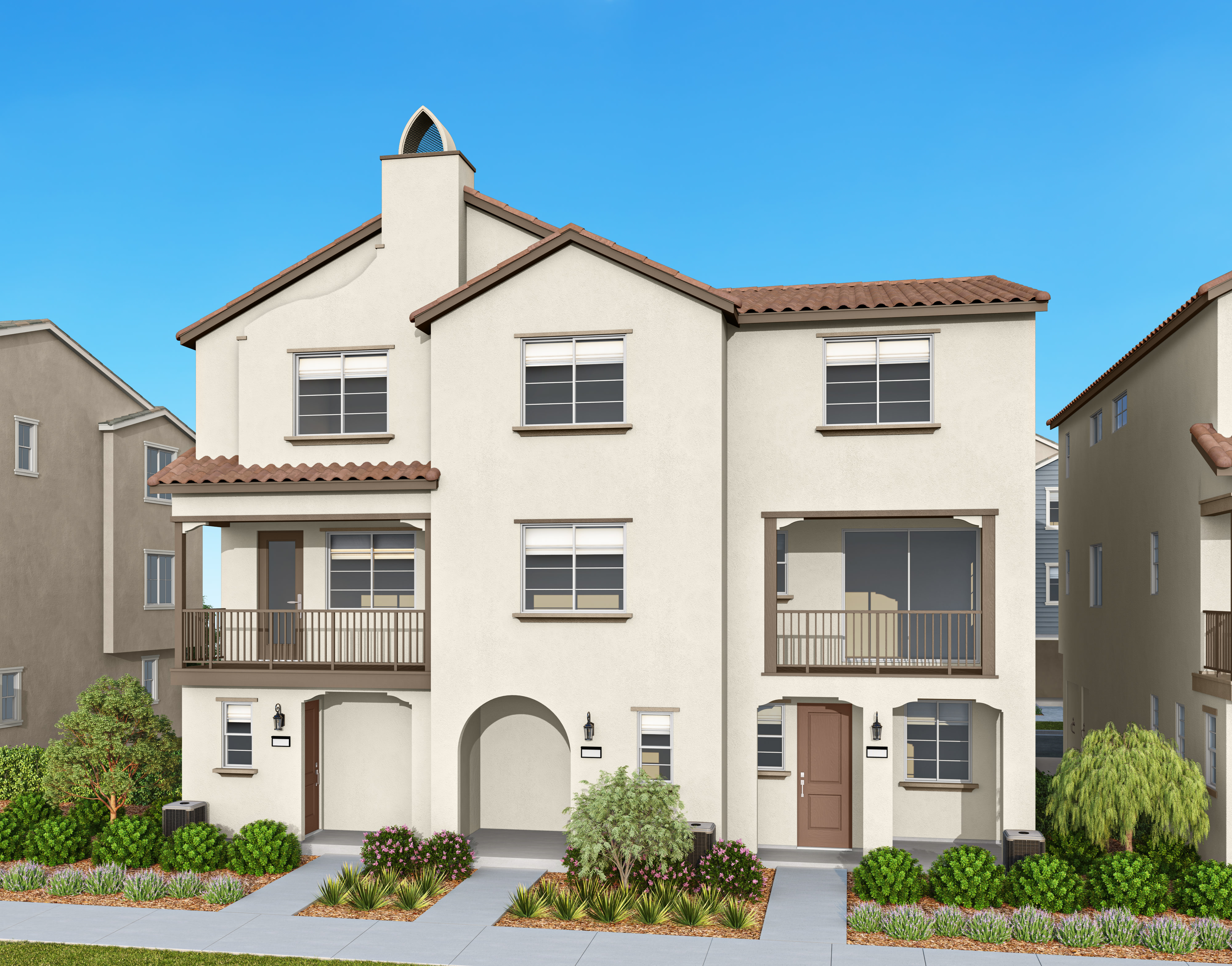 A two-story residential building with a Mediterranean-style architecture, featuring a tiled roof, arched windows, and a balcony, set against a clear blue sky and surrounded by lush greenery.