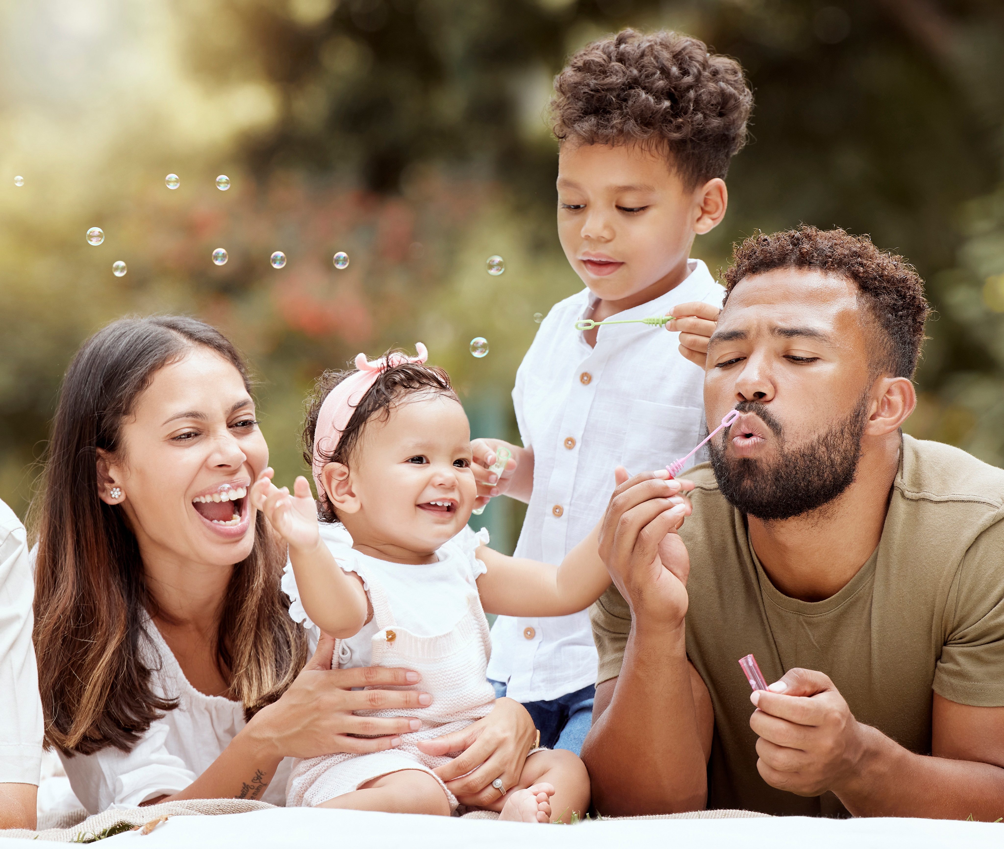 A happy, diverse family sitting together outdoors, surrounded by a blurred natural setting with sunlight filtering through.