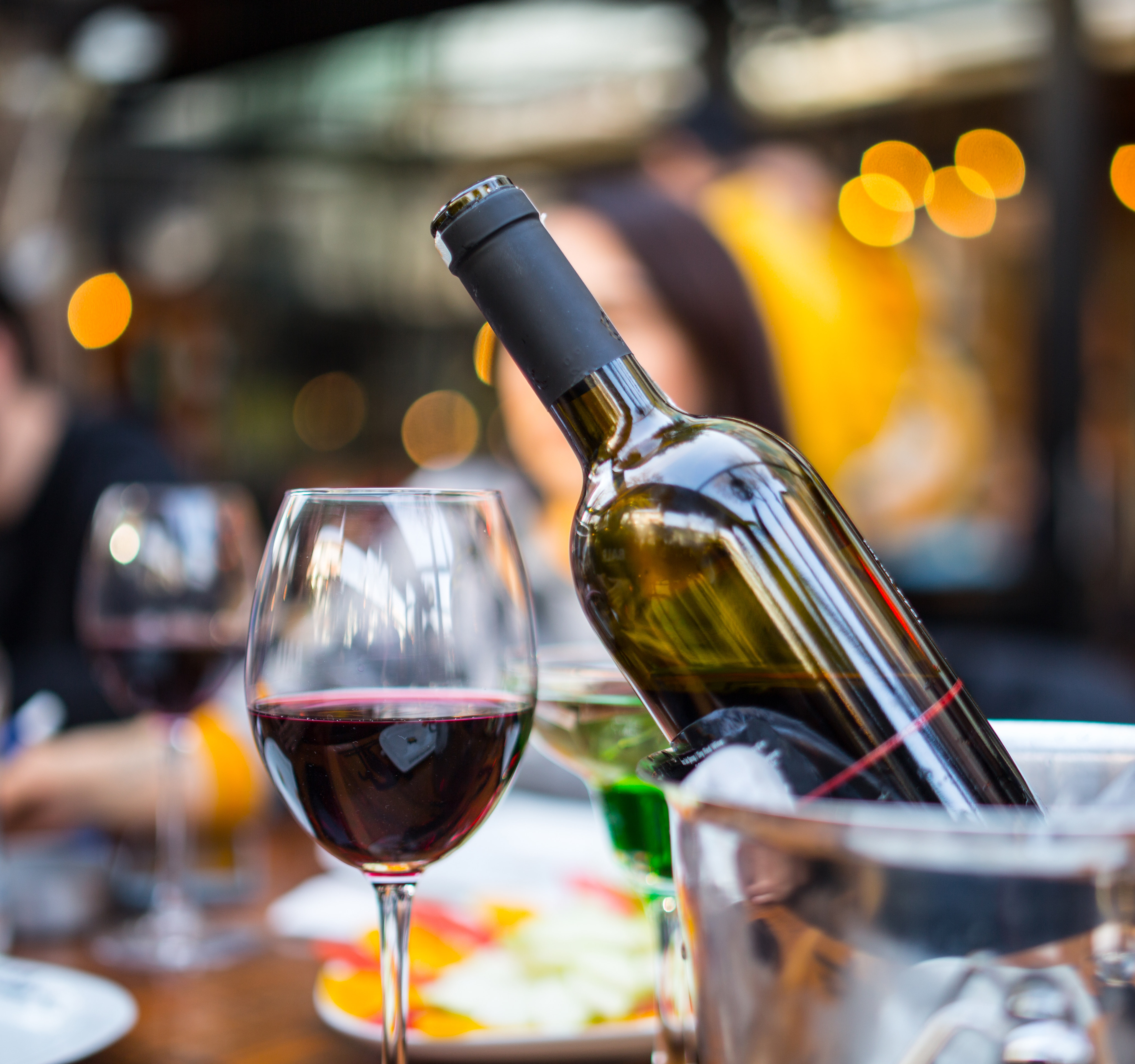 A bottle of wine and a glass of red wine are prominently displayed in the foreground, with a blurred background of what appears to be a restaurant or bar setting with colorful lights.