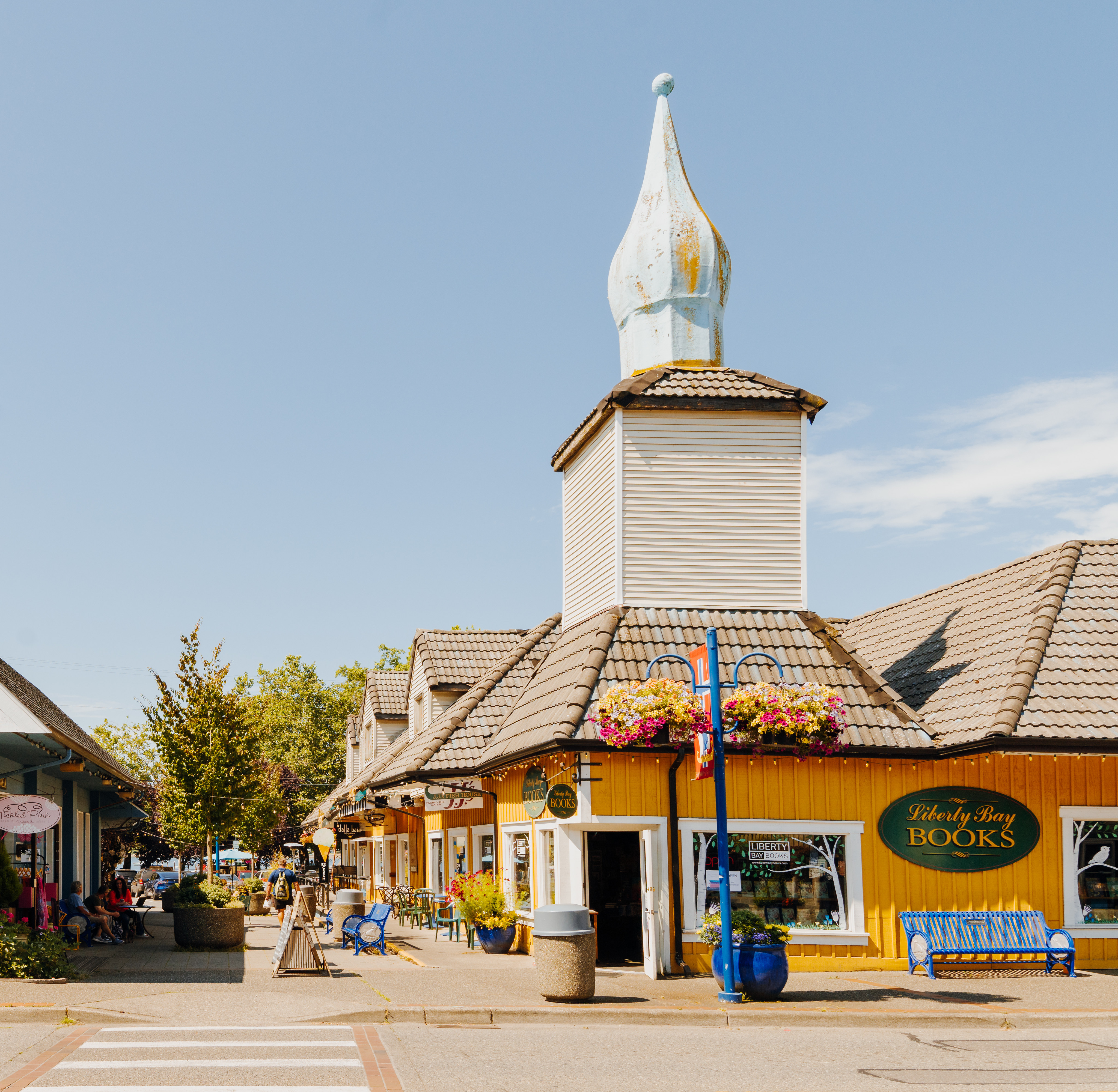 A quaint and colorful village street with a distinctive spire-topped building, surrounded by shops and pedestrians, set against a clear blue sky.