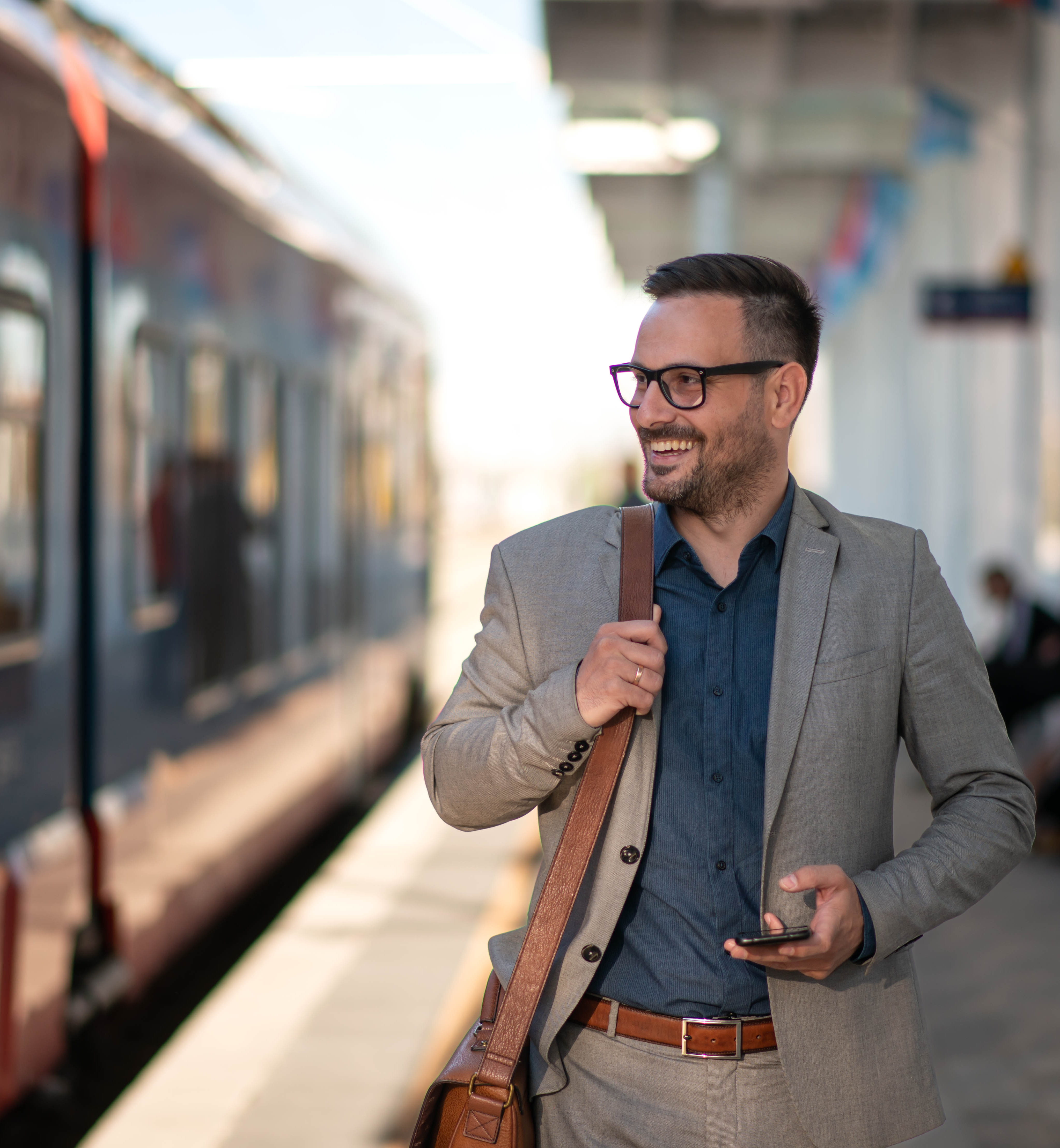 A man in a gray suit and glasses stands on a train platform, holding a smartphone and smiling.